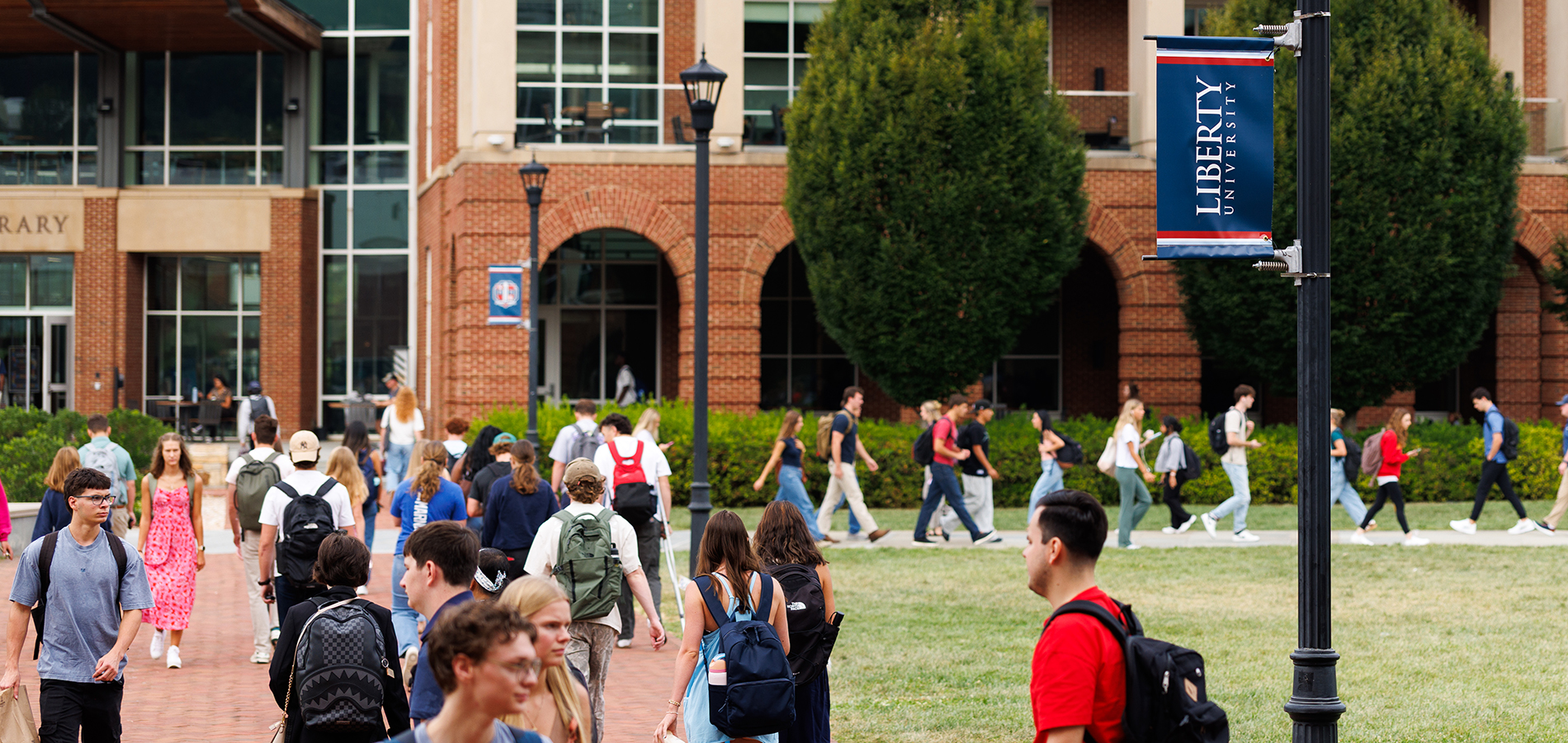 Students walk across a campus lawn in front of a brick building with a "Liberty University" banner on a lamppost.