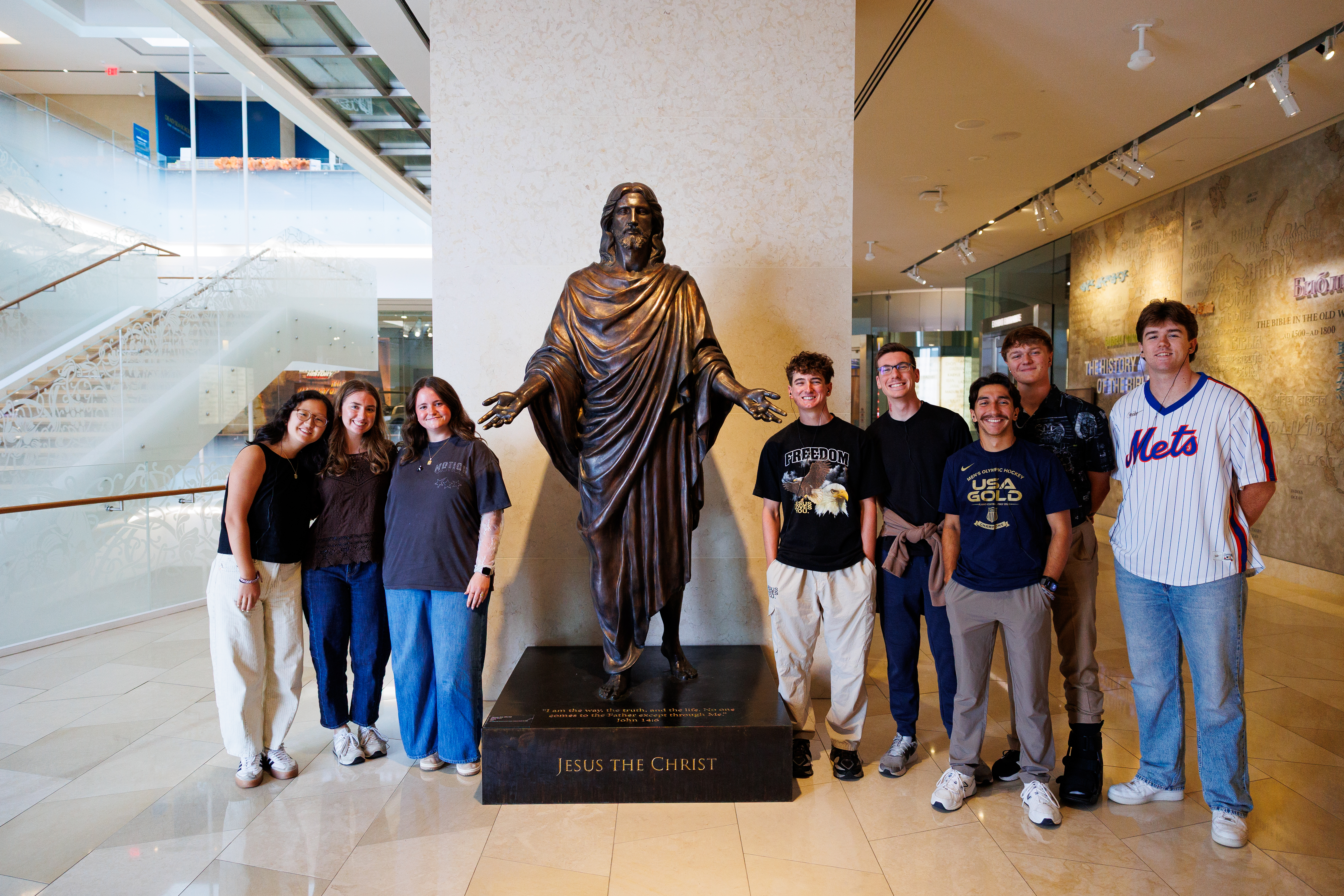 A group of young people poses for a photo beside a large bronze statue of Jesus the Christ in a modern indoor setting.