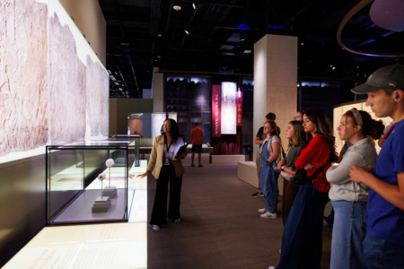 A guide stands before a group of visitors in a museum exhibit, gesturing toward a display case containing small artifacts under bright lighting.