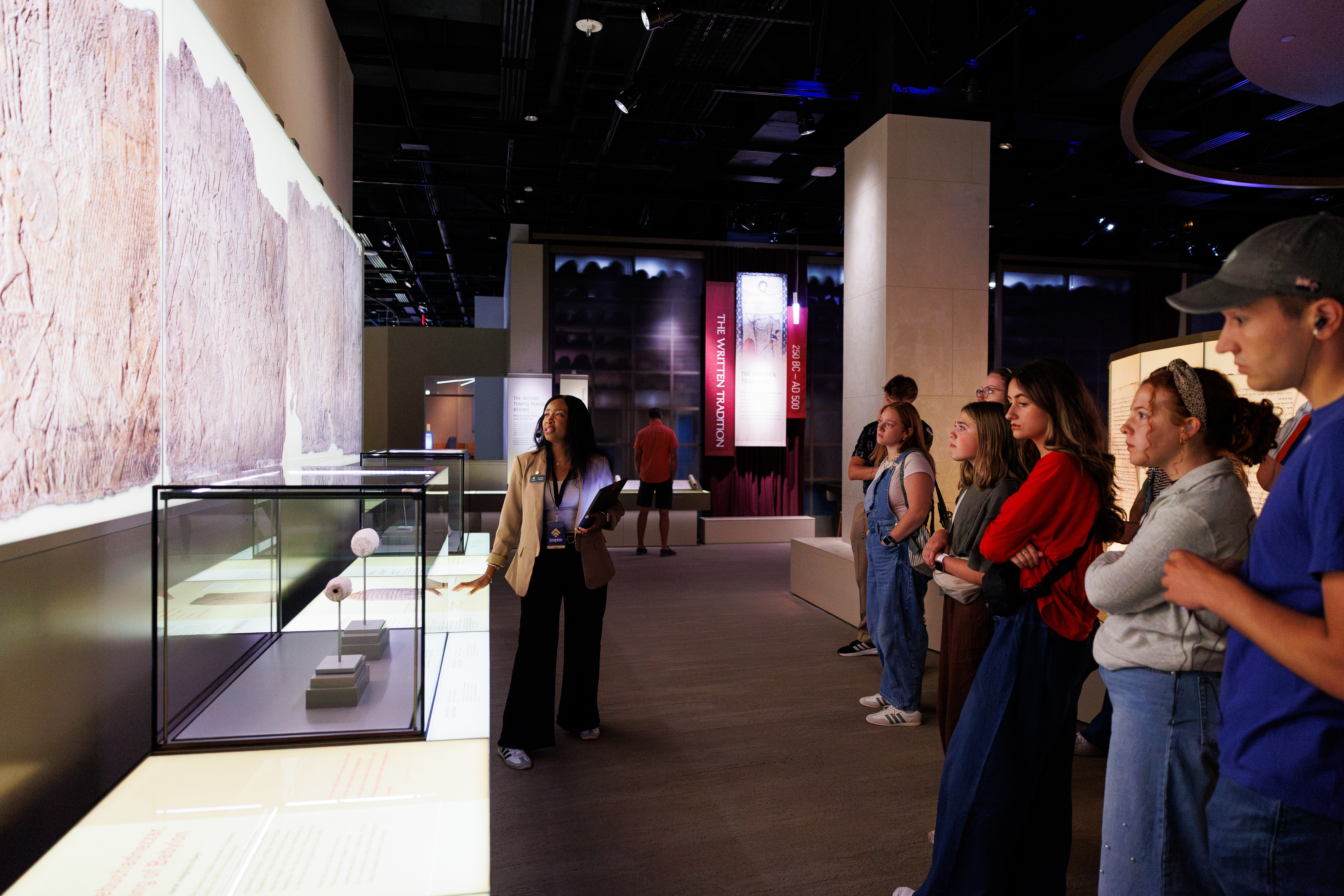 A guide stands before a group of visitors in a museum exhibit, gesturing toward a display case containing small artifacts under bright lighting.