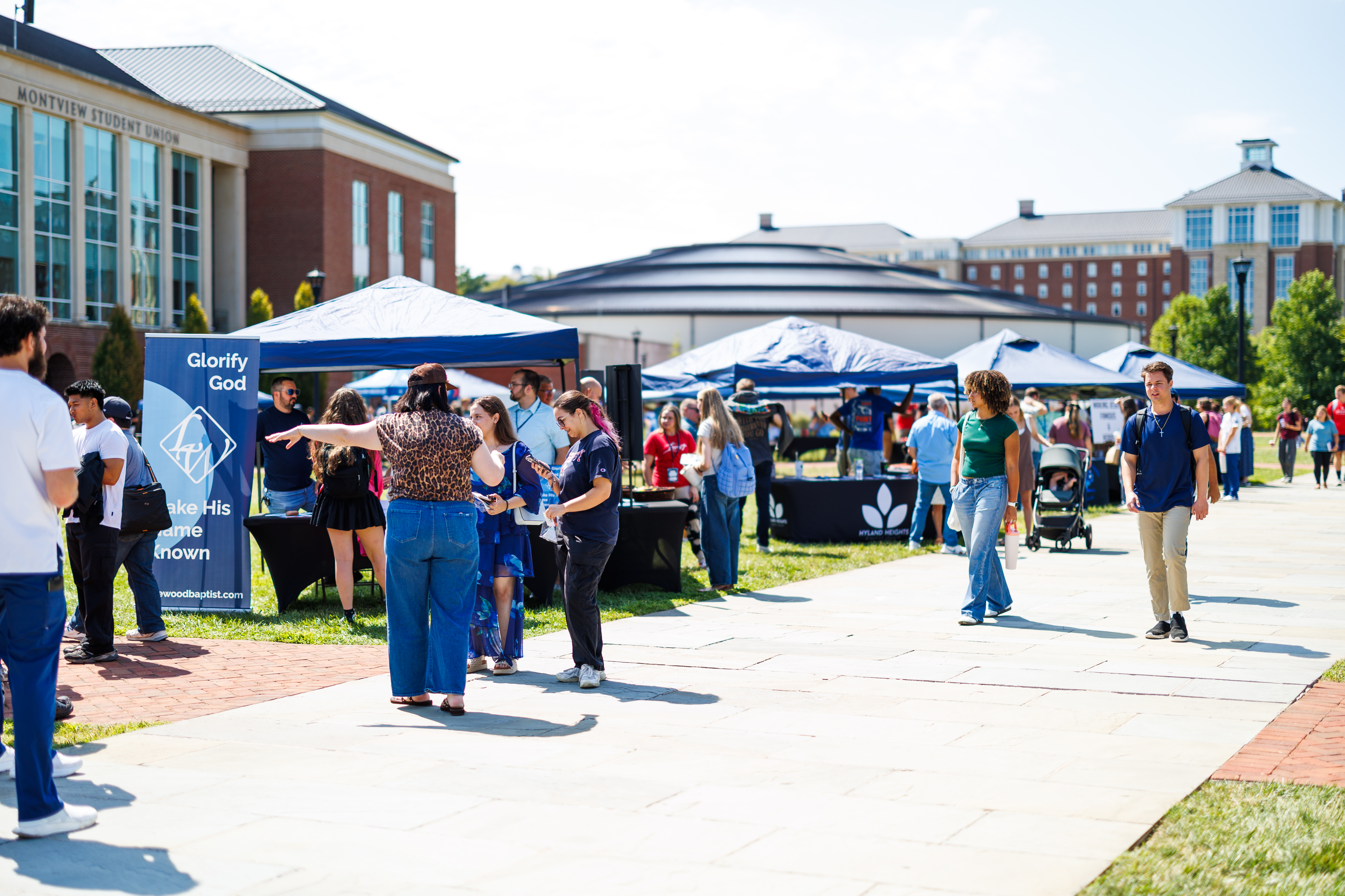 A group of people gather under blue tents on a sunny day at a campus event near the Montview Student Union building.