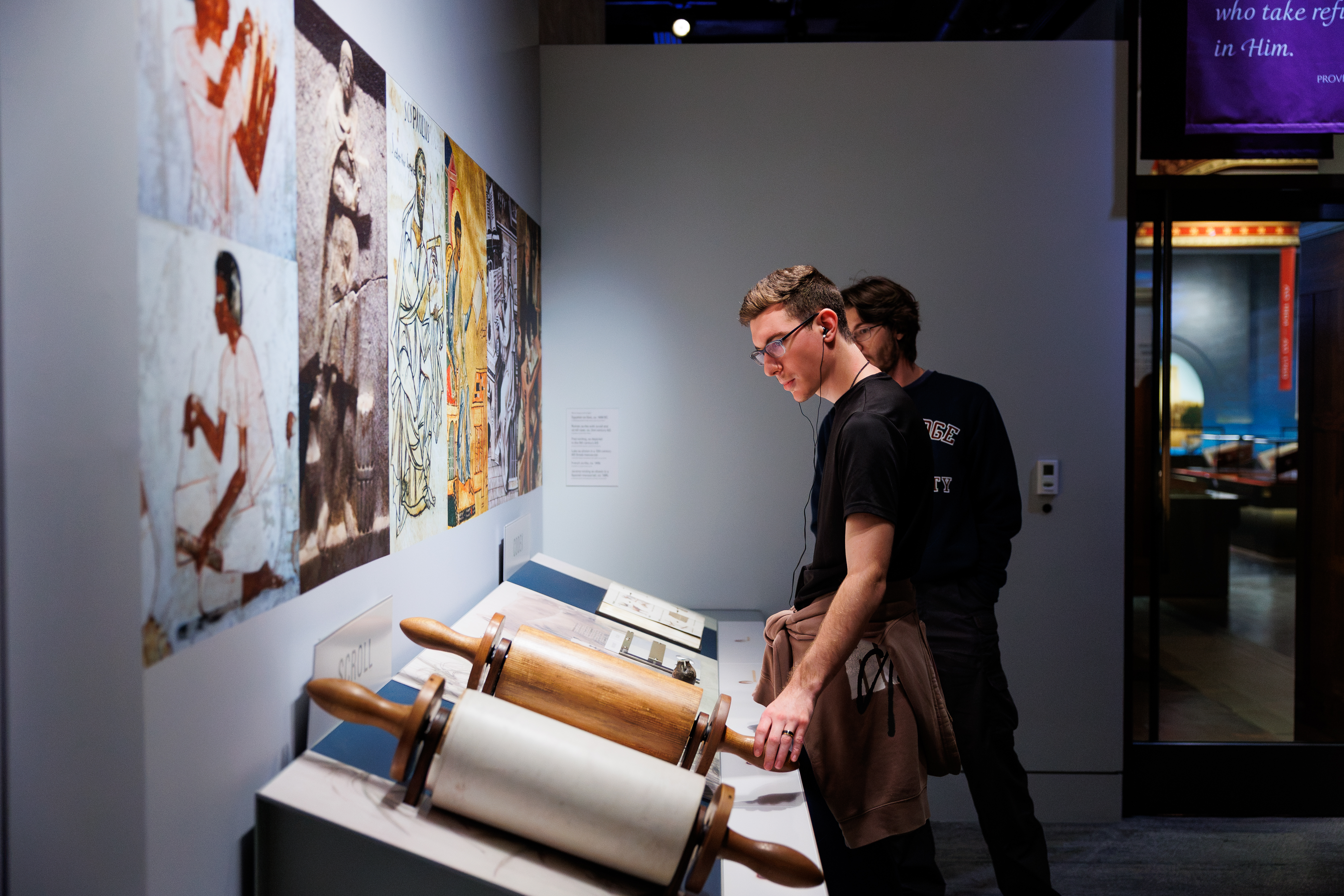 Two individuals examine historical artifacts, including wooden rollers, displayed in a museum exhibit.