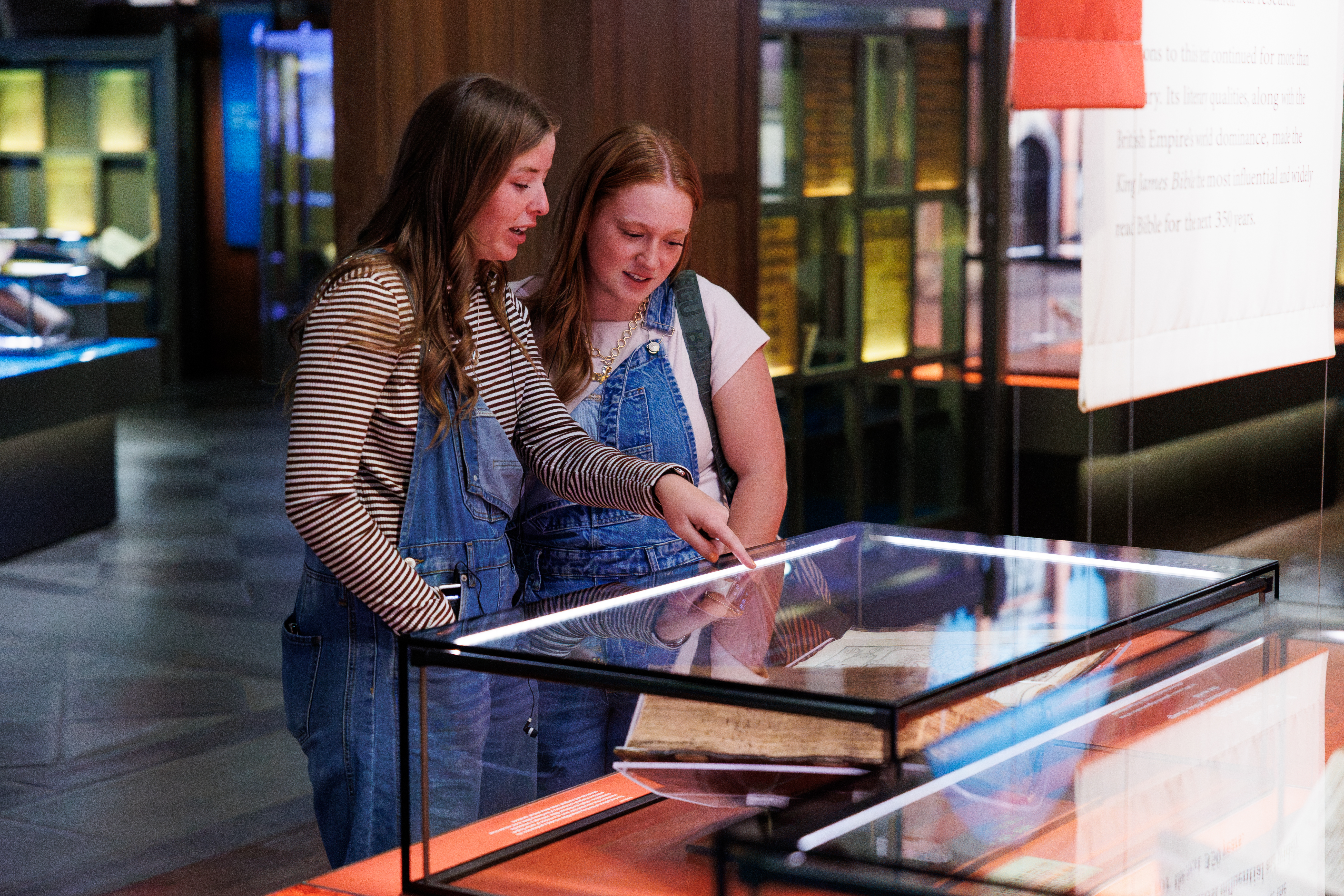 Two individuals examine a historical book displayed in a glass case within a museum setting.