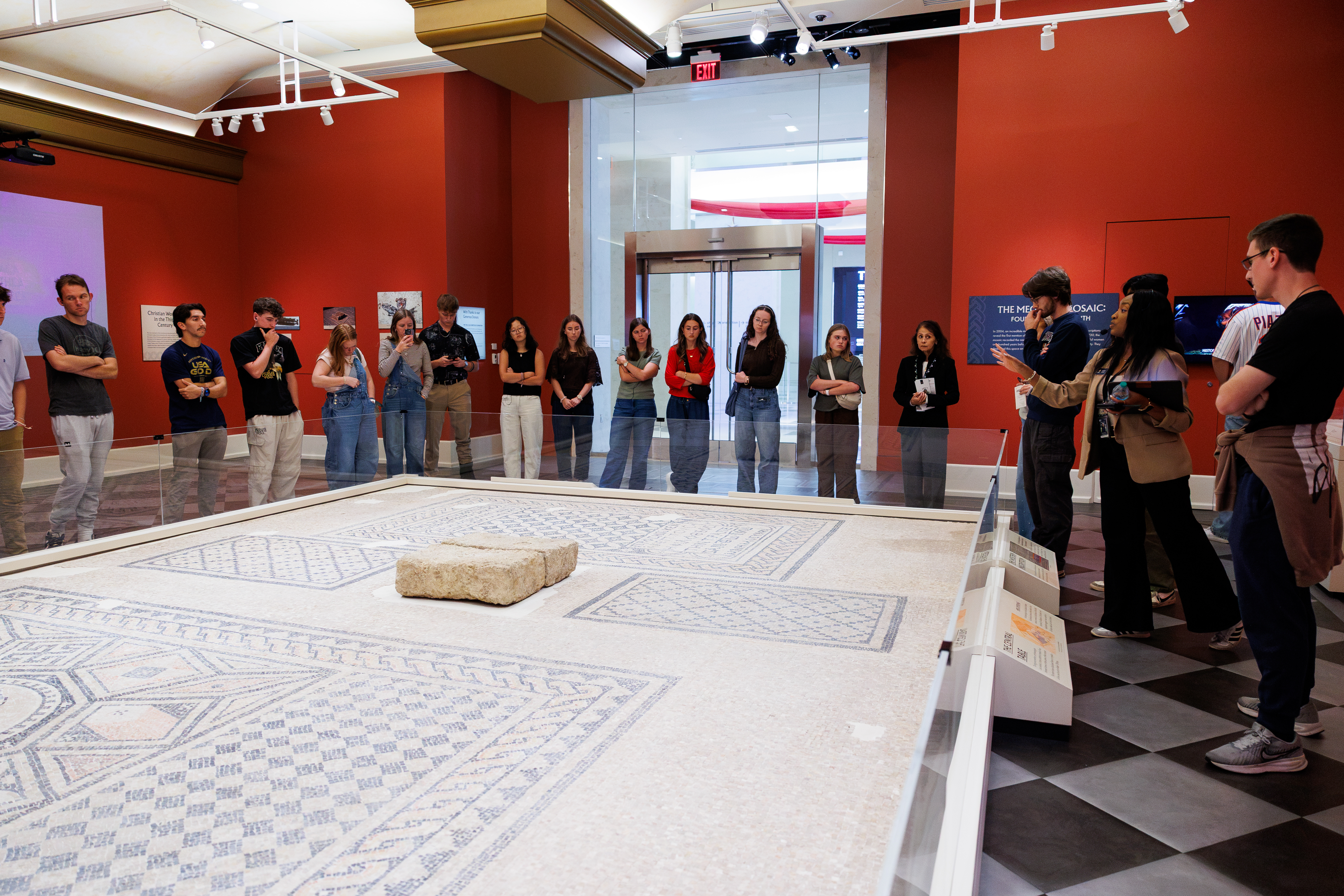 A group of visitors observes a large ancient mosaic displayed in a museum exhibit with red walls and modern lighting.