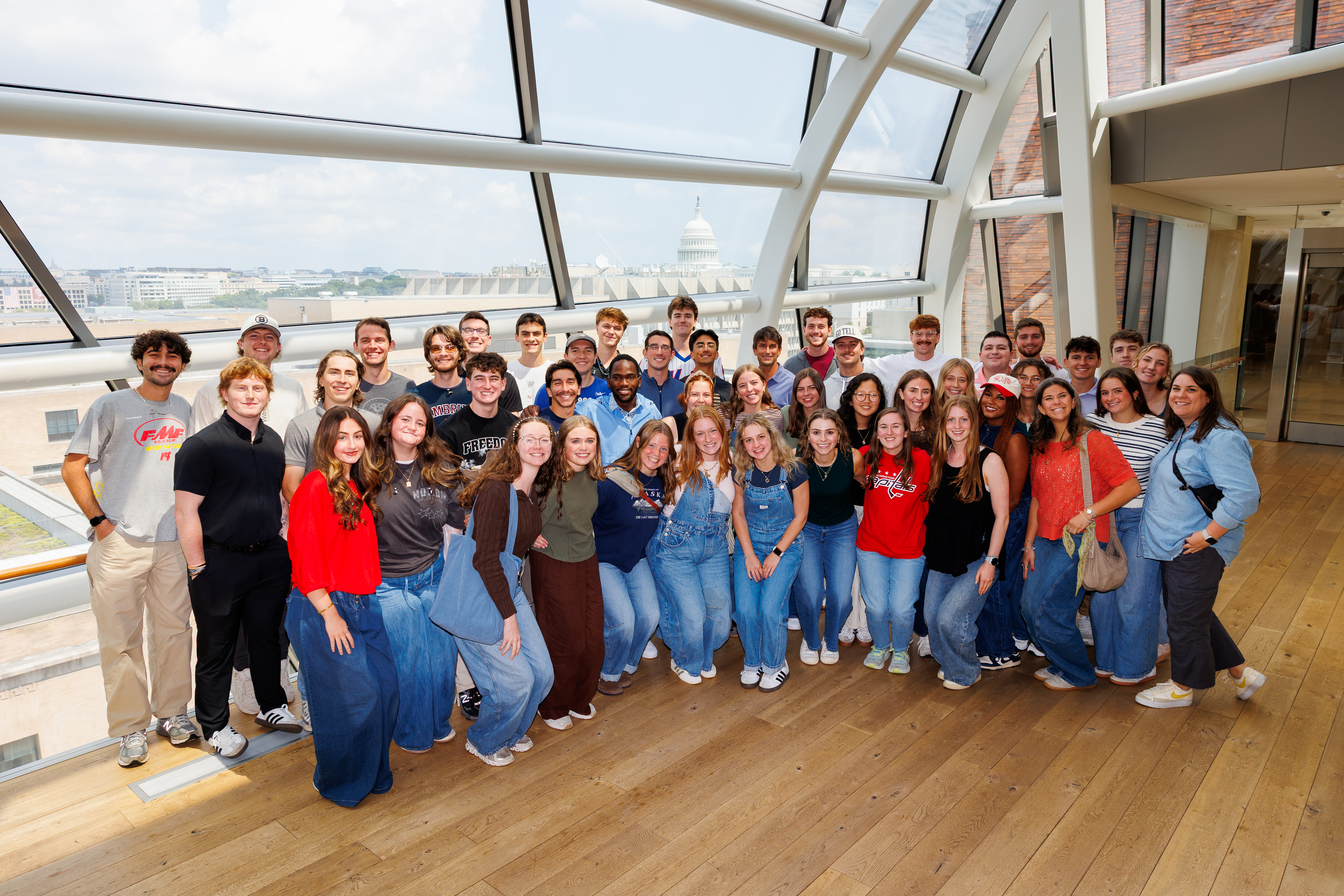 A group of young people pose together indoors near a large window overlooking a cityscape with a prominent capitol building in the distance.