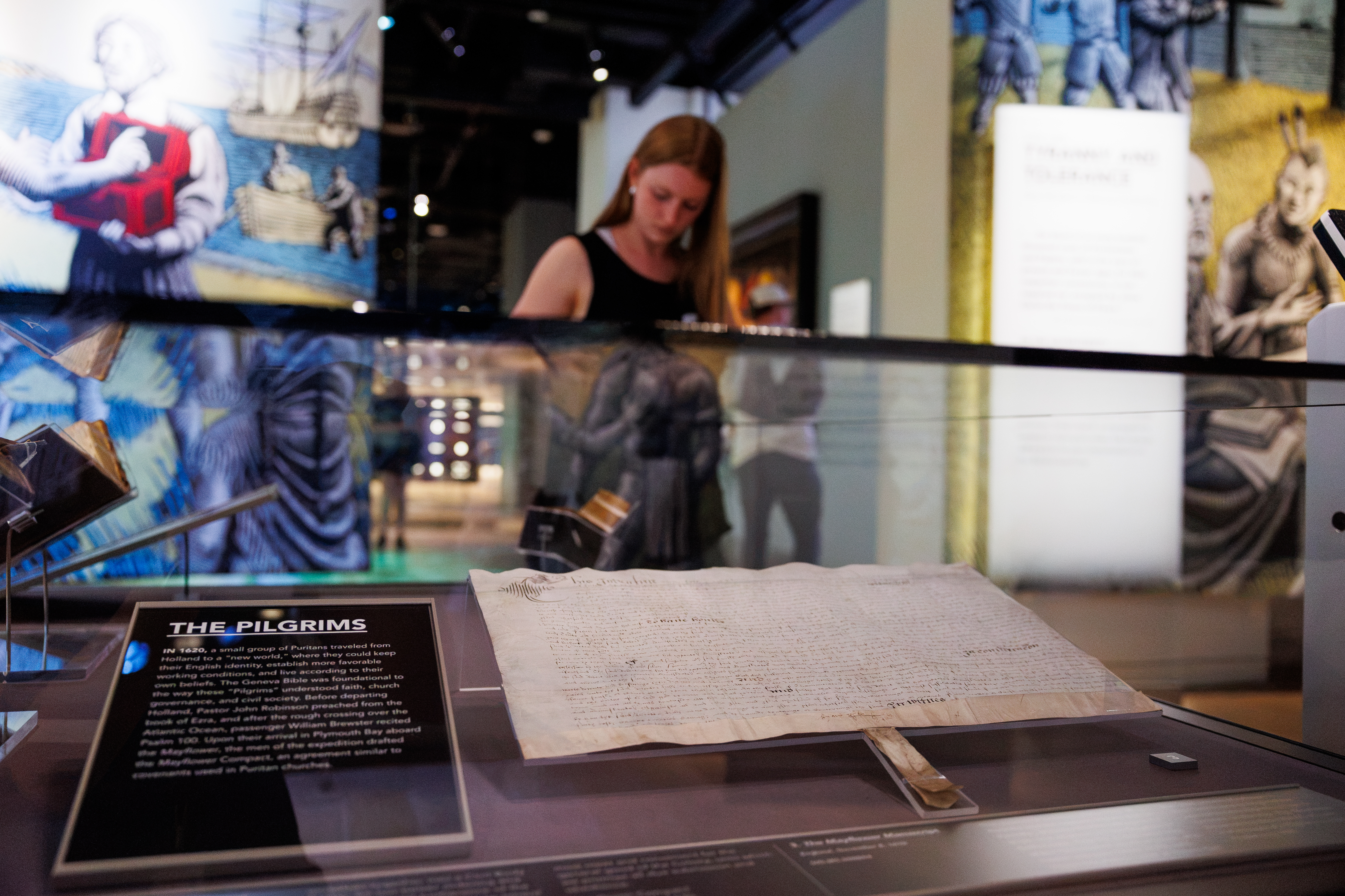 A historical document displayed in a museum exhibit, accompanied by an informational plaque about the Pilgrims.