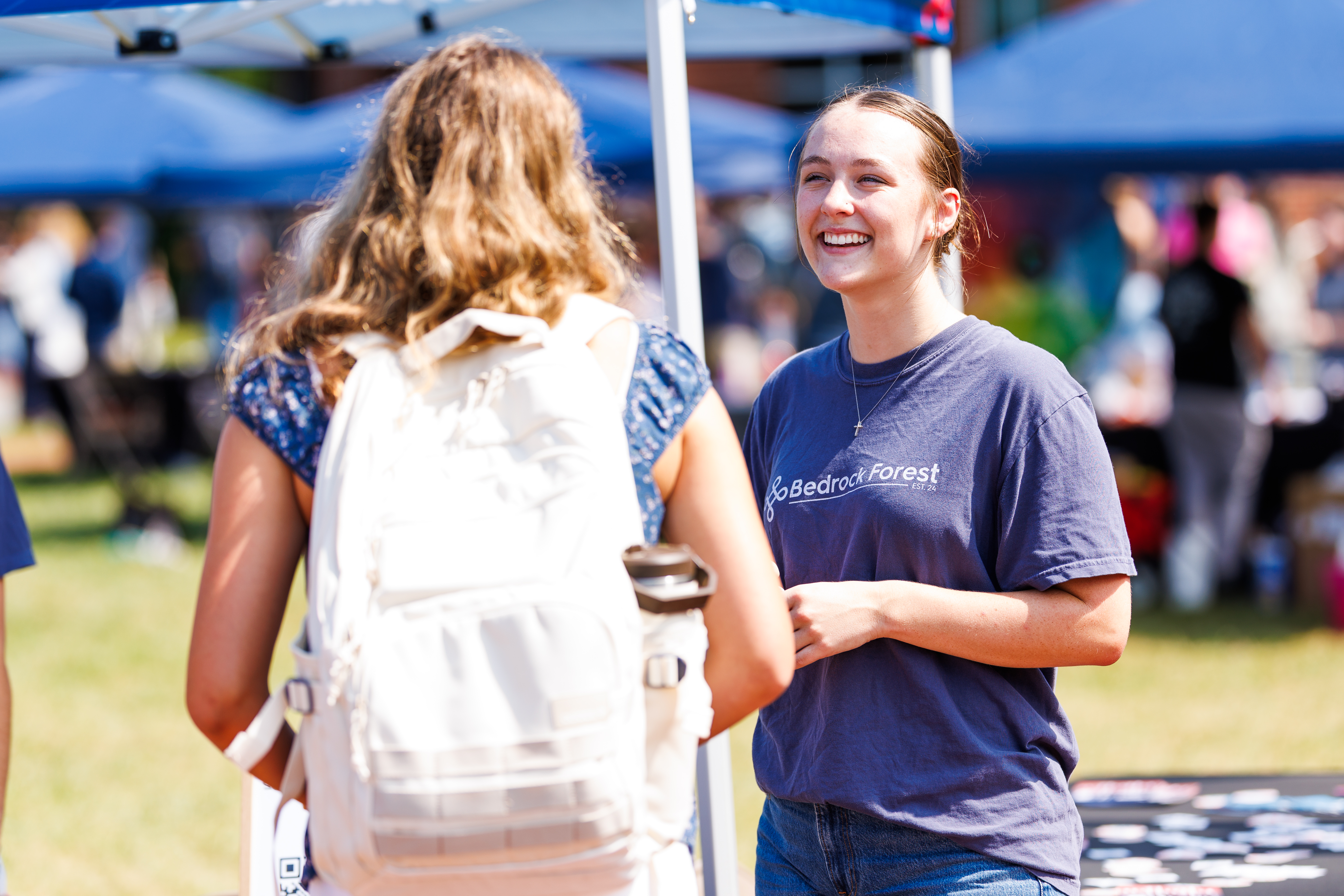 Two individuals engage in conversation at an outdoor event under blue tents, one wearing a "Bedrock Forest" shirt and carrying a white backpack.