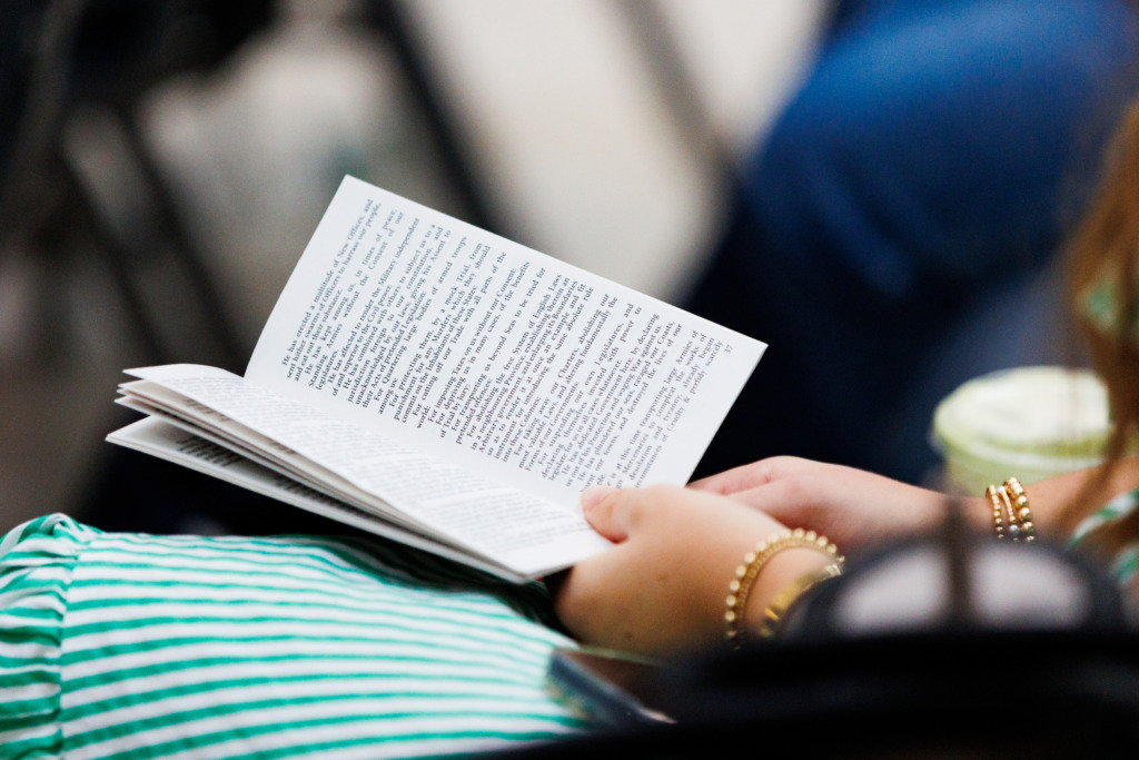A person in a green and white striped shirt holds an open book while seated, with hands adorned with bracelets.