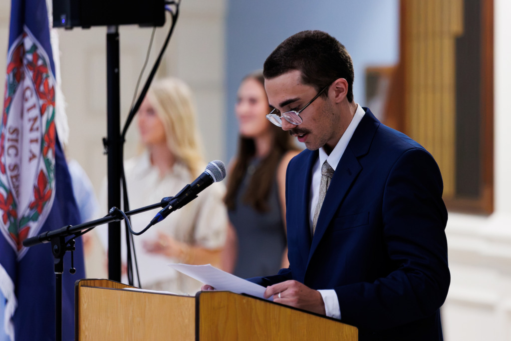 A person in a suit stands at a podium, reading from papers while speaking into a microphone, with a flag and blurred figures in the background.