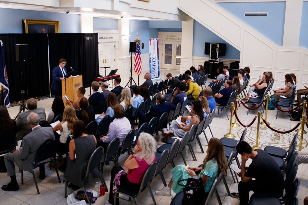 A person speaks at a podium before an audience seated in rows within a formal hall.
