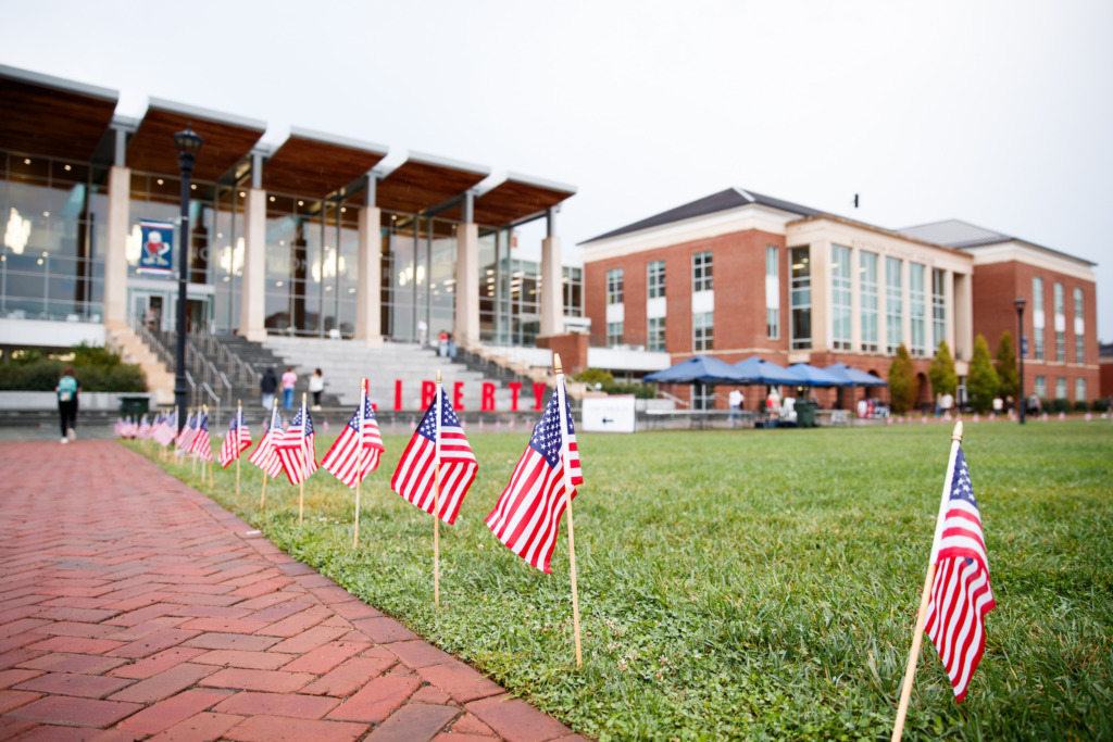 A row of American flags lines a grassy area beside a brick walkway, with modern and brick buildings in the background.