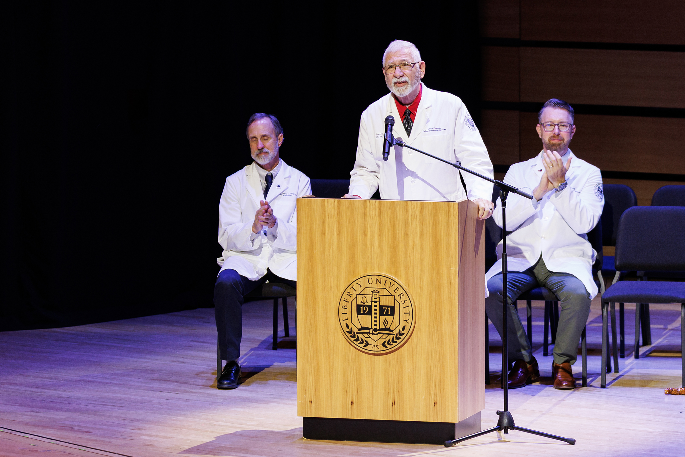 A person in a white coat speaks at a podium on a stage, with others in white coats seated nearby, applauding.