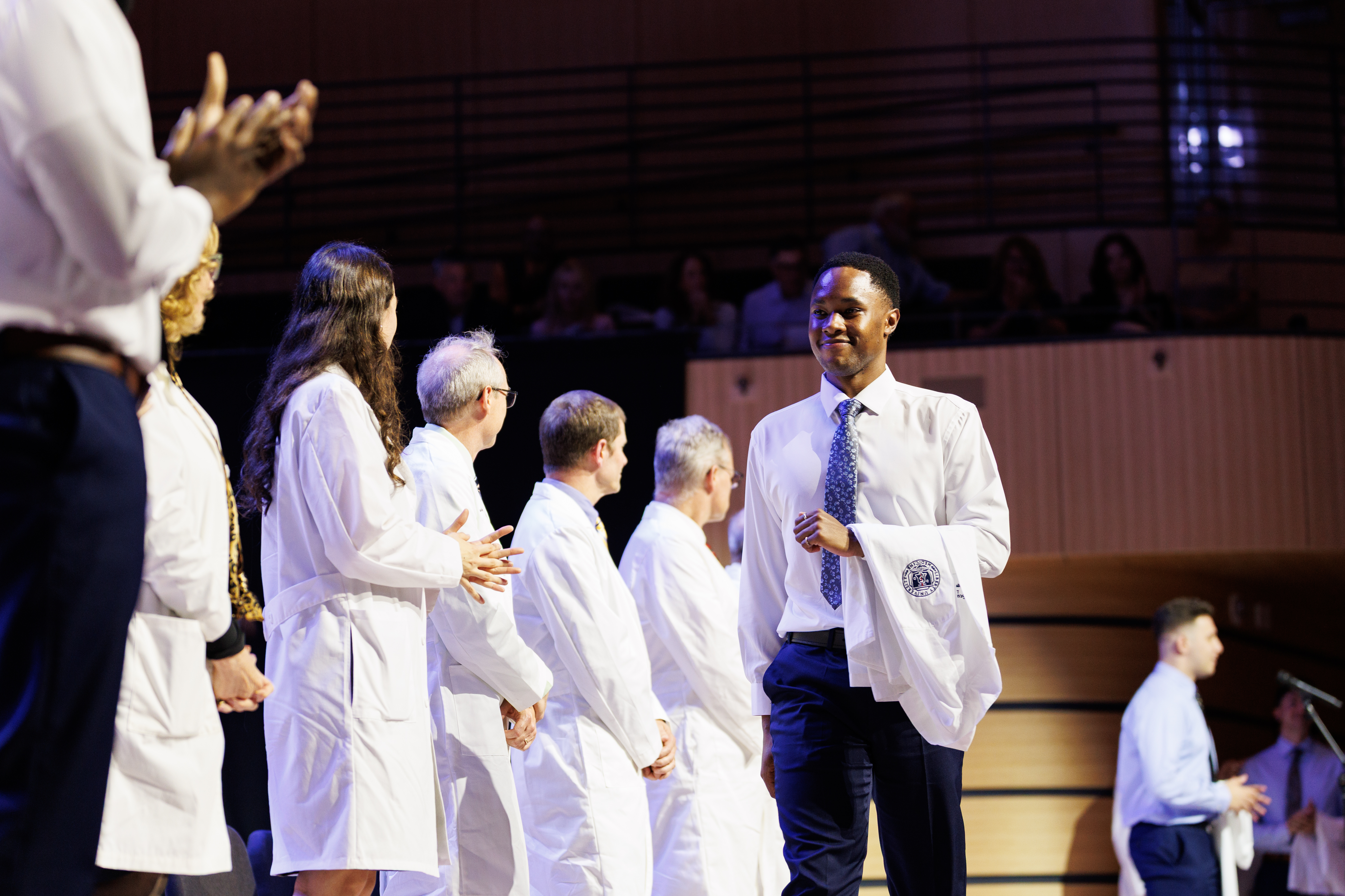A group of individuals in white coats stand on a stage, with one person smiling and holding a coat, as others clap and observe.