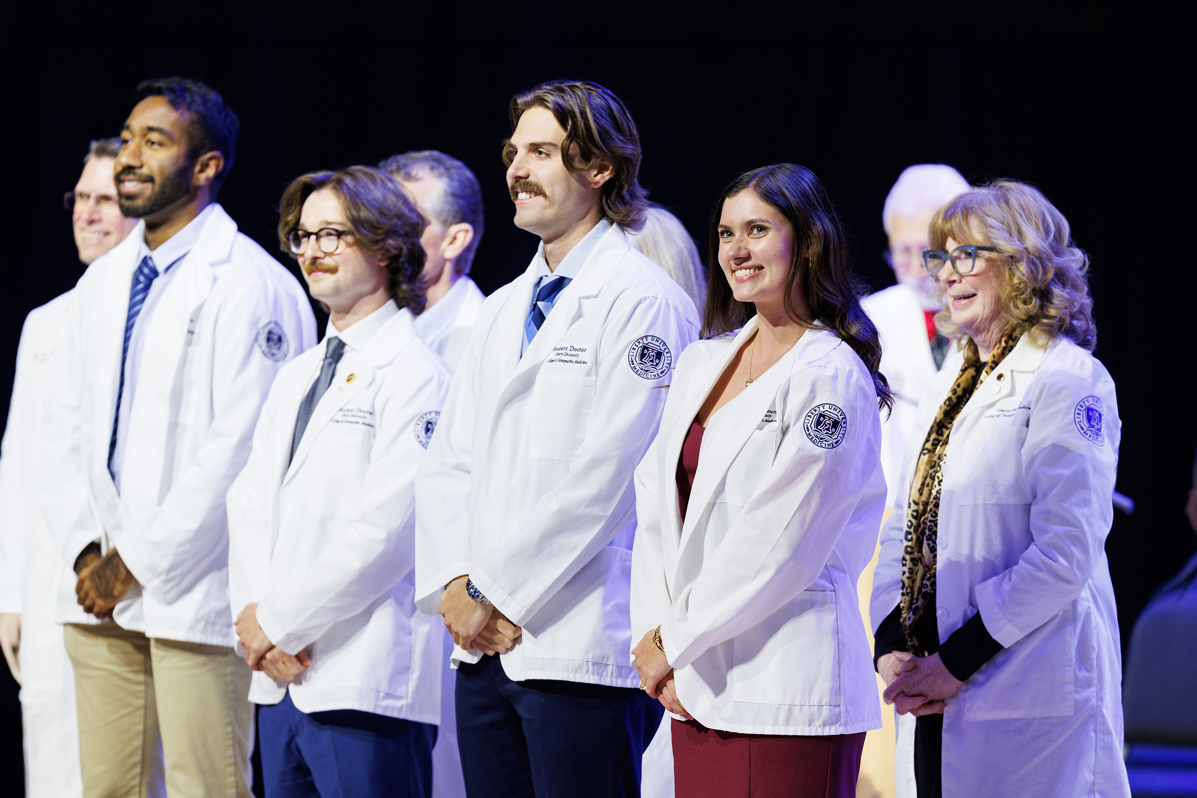 A group of individuals in white lab coats stand together on a stage, smiling and looking toward the right.
