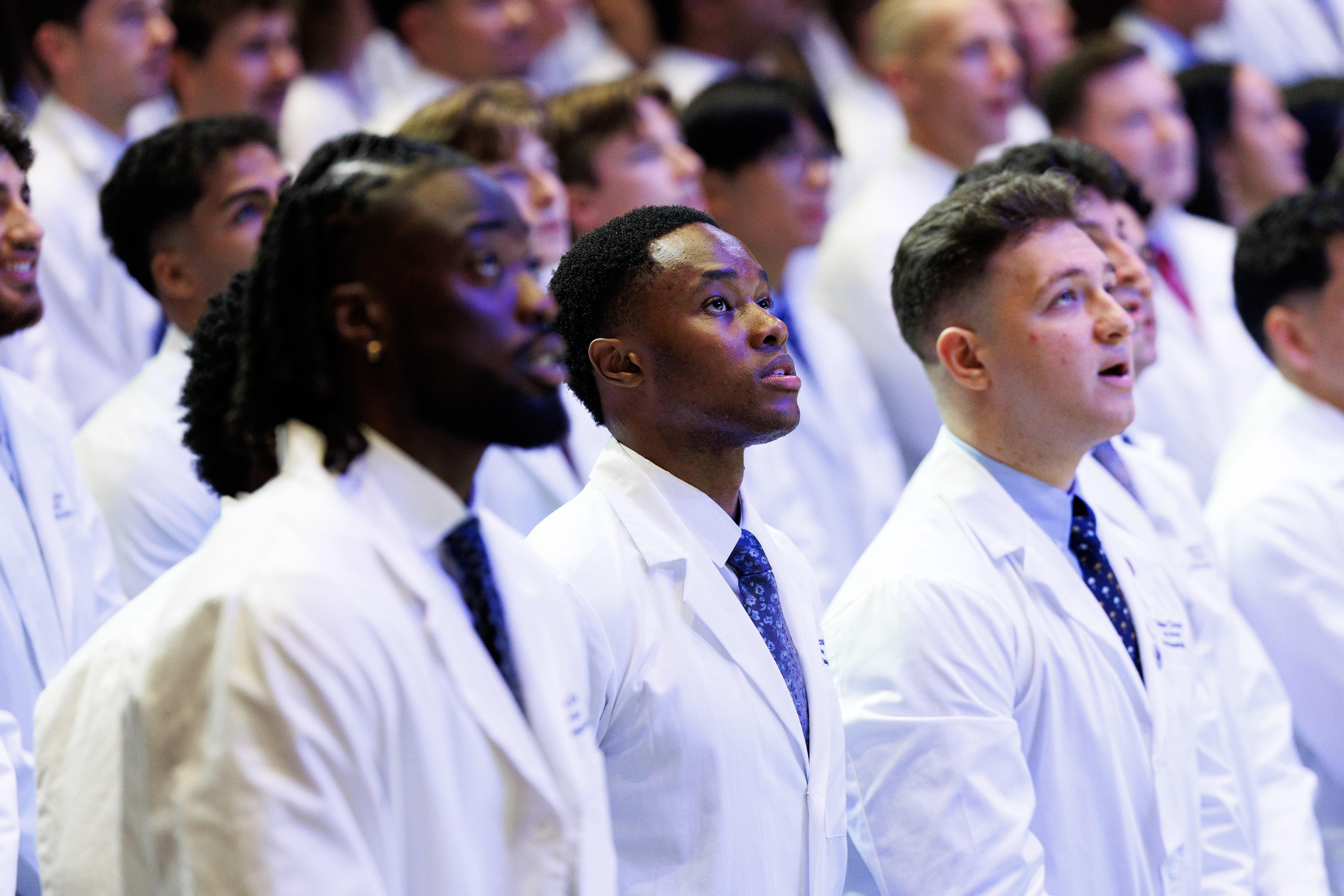 A group of individuals in white lab coats stand attentively, looking upward during what appears to be a formal ceremony or event.