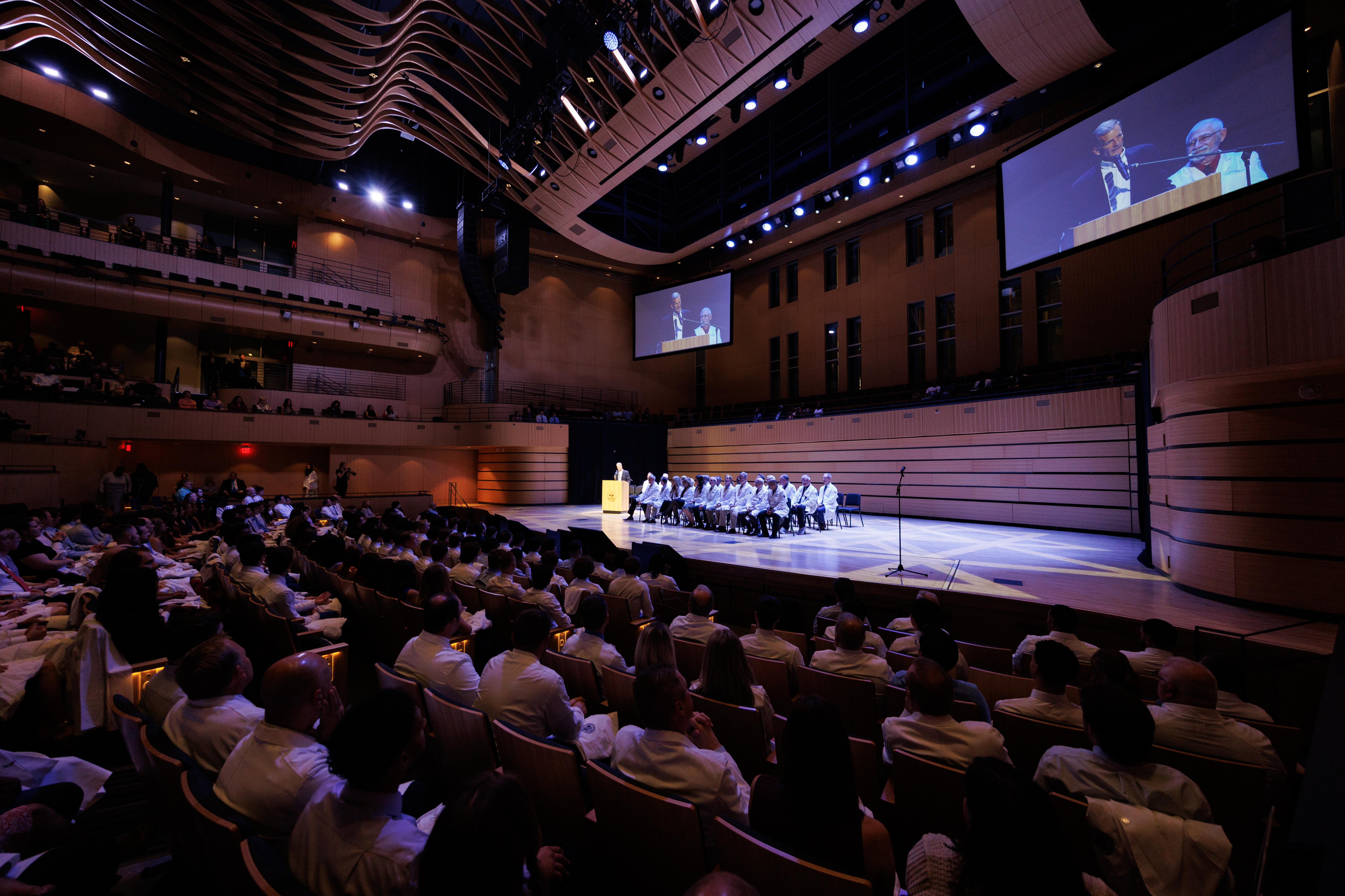 A large auditorium filled with attendees watches a formal event on stage, where a group in white attire sits while two individuals speak, projected on screens.