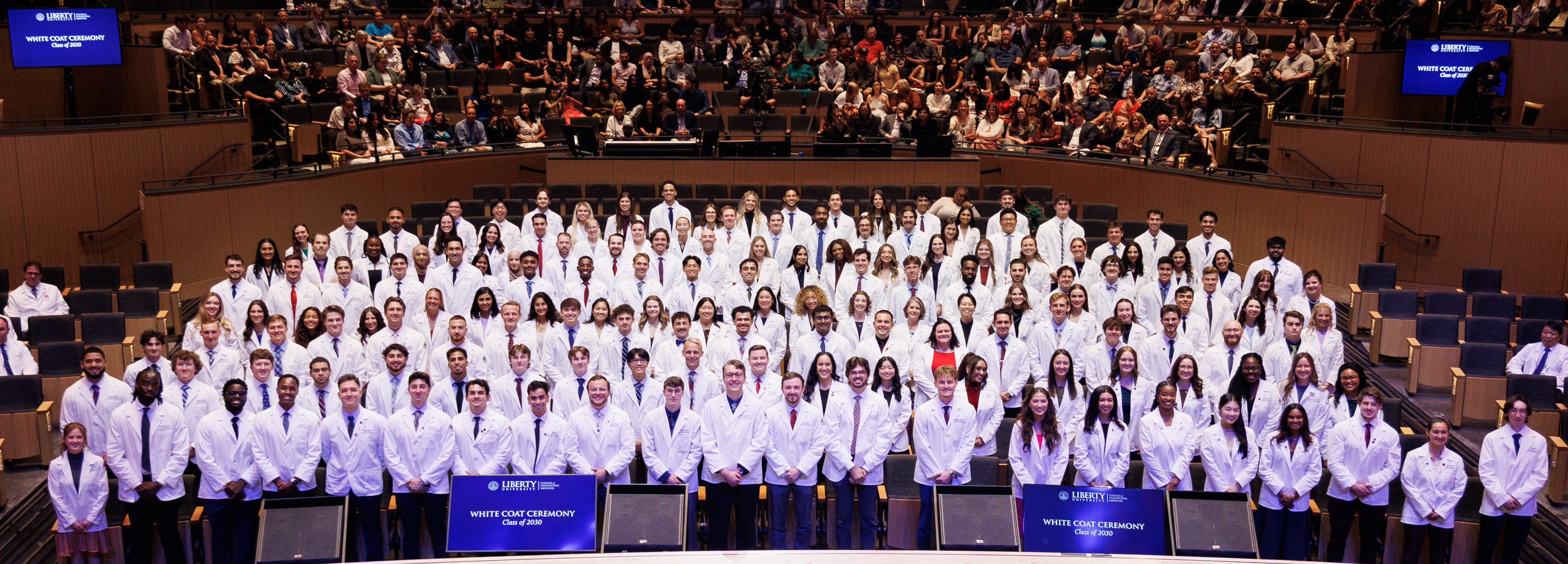 A large group of individuals in white coats poses for a photo during a White Coat Ceremony at Liberty University, with an audience seated in the background.