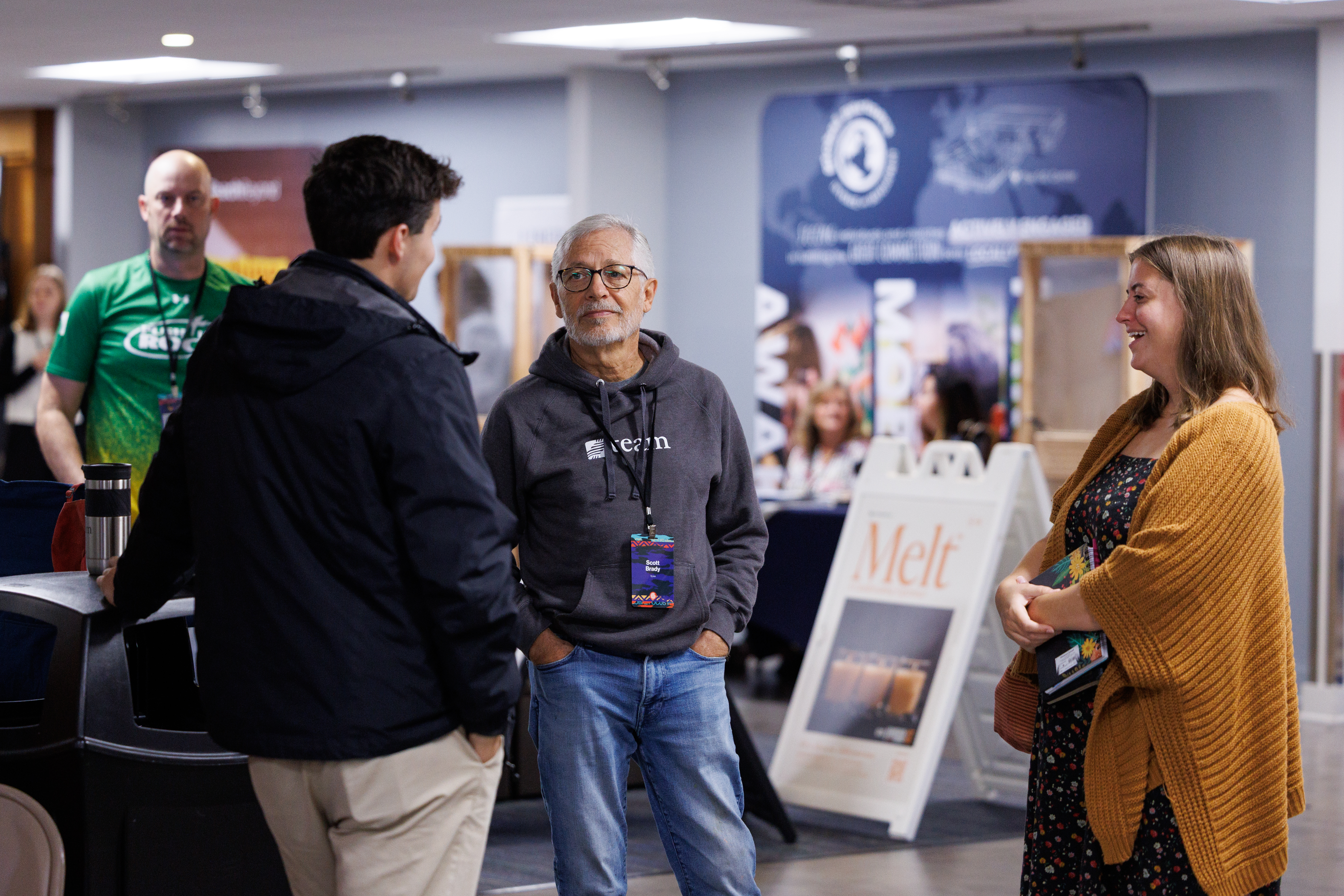 A group of individuals engages in conversation at an indoor event, with one person smiling while holding a folder and another wearing a hoodie and glasses.