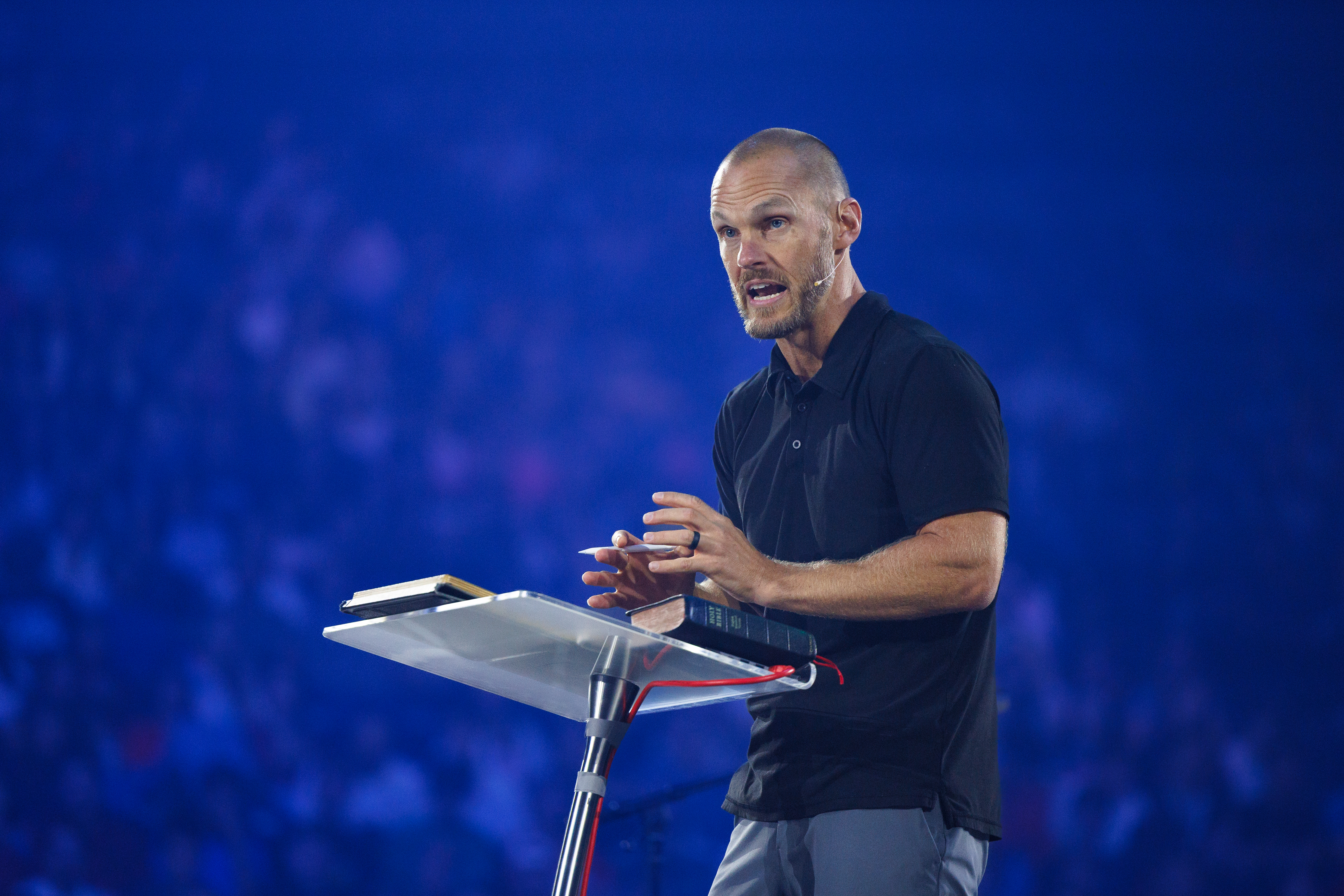 A person stands at a podium, speaking into a microphone while holding a book, against a blurred blue background.