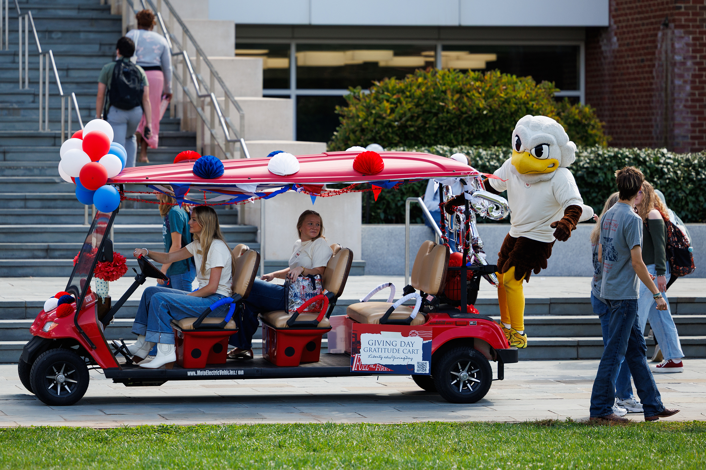A red golf cart decorated with balloons and flags, featuring a mascot and passengers, moves through a campus setting.