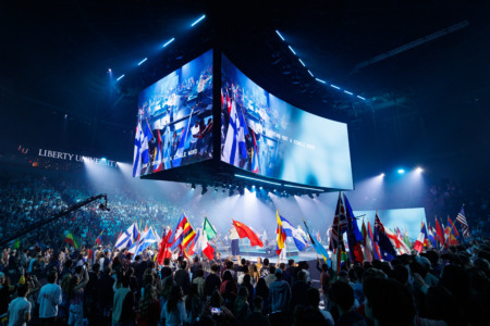 A large crowd gathers in a dimly lit arena at Liberty University, waving numerous international flags beneath a massive suspended screen displaying a live event.