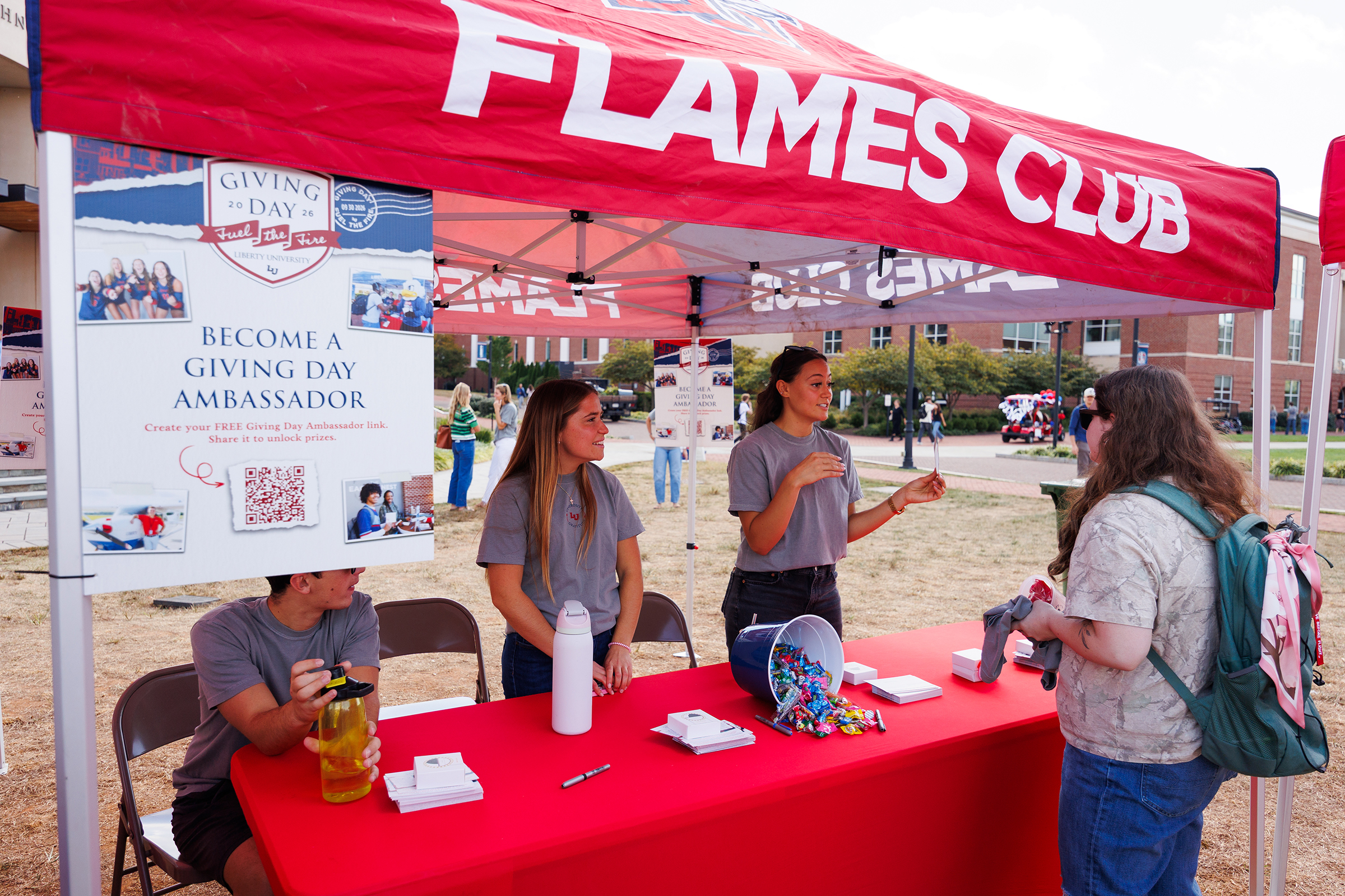 Three individuals engage at a red-tented Flames Club booth during Giving Day, where one person interacts with a visitor while another sips a drink.