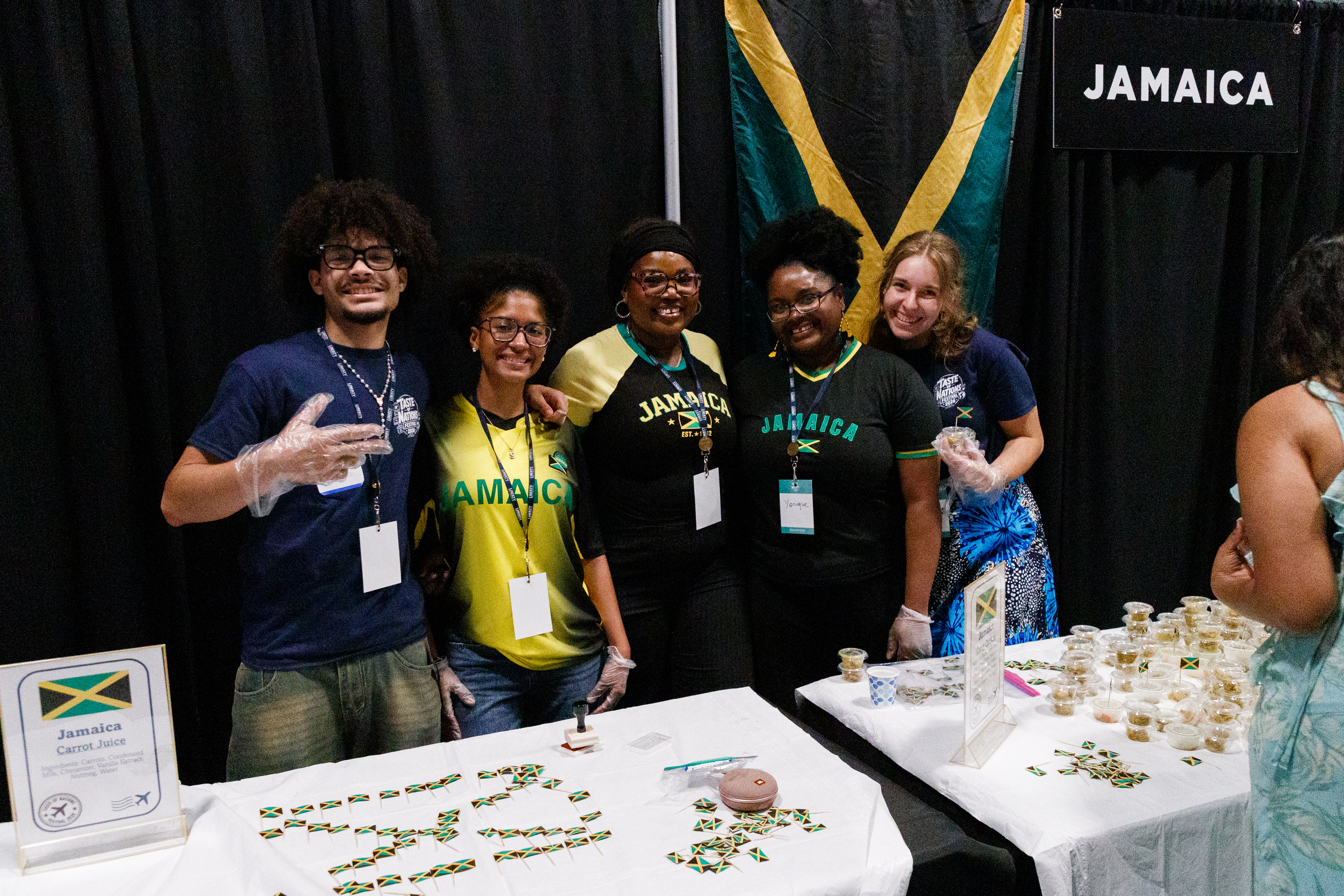 A group of five individuals in Jamaica-themed attire stand behind tables displaying Jamaican flag-themed items and carrot juice at a booth with a Jamaican flag backdrop.