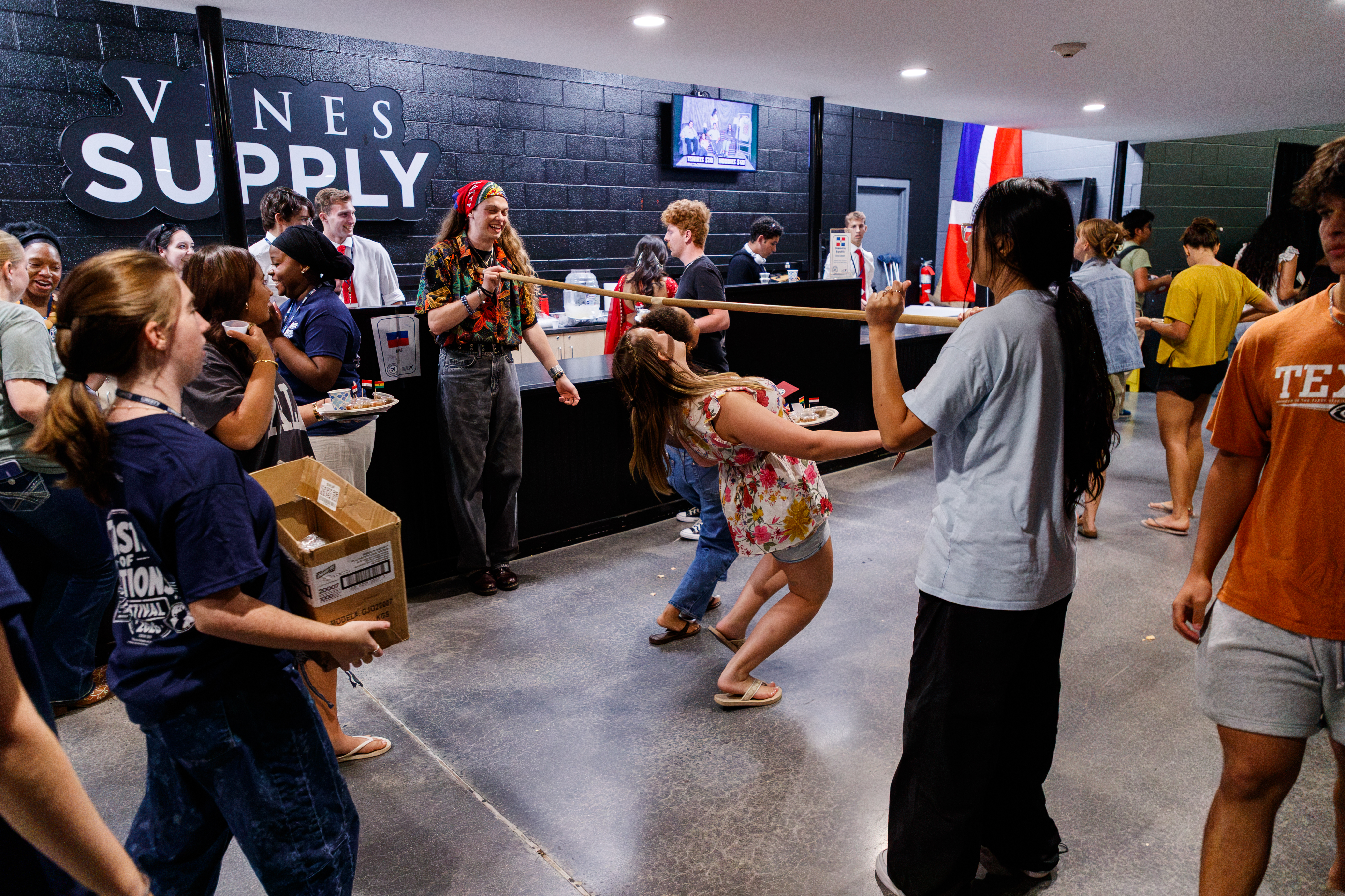A group of people gather in a store called "Vines Supply," where some are playing a game with a long stick while others watch and socialize.
