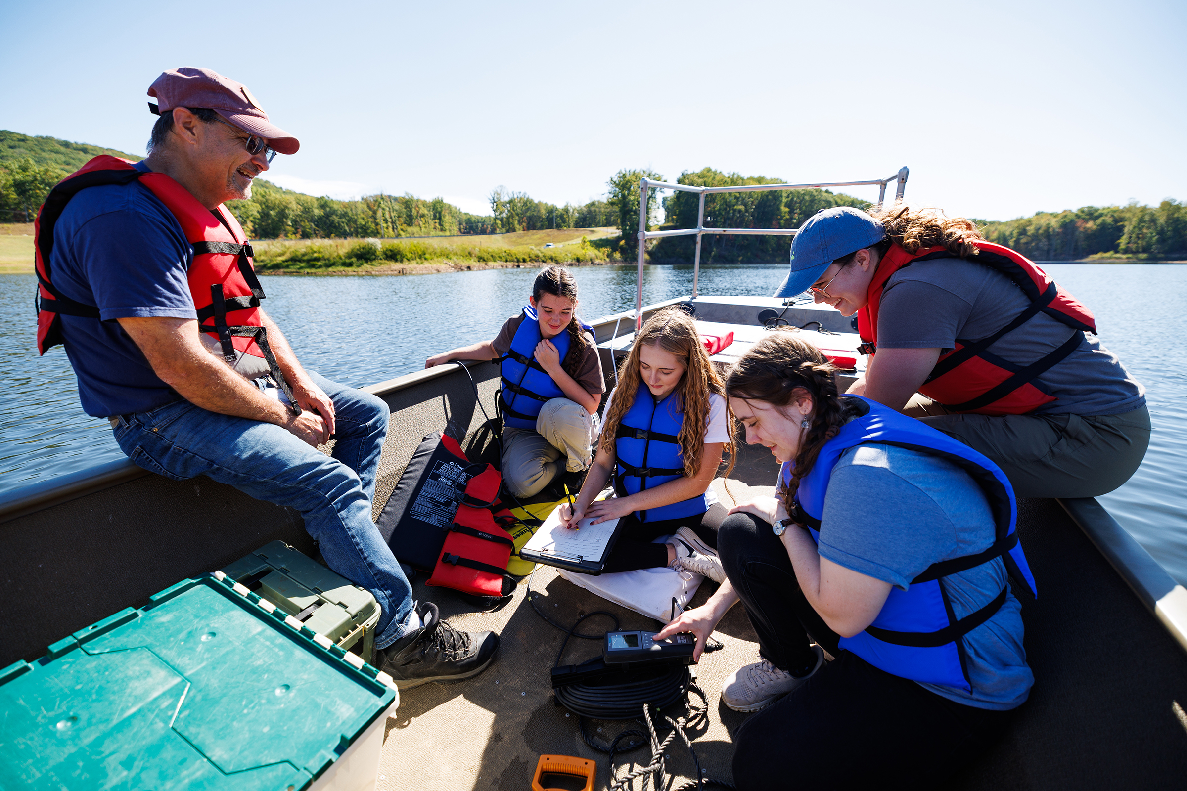 A group of people in life jackets on a boat, with some writing notes and others observing, while a man smiles from the side.