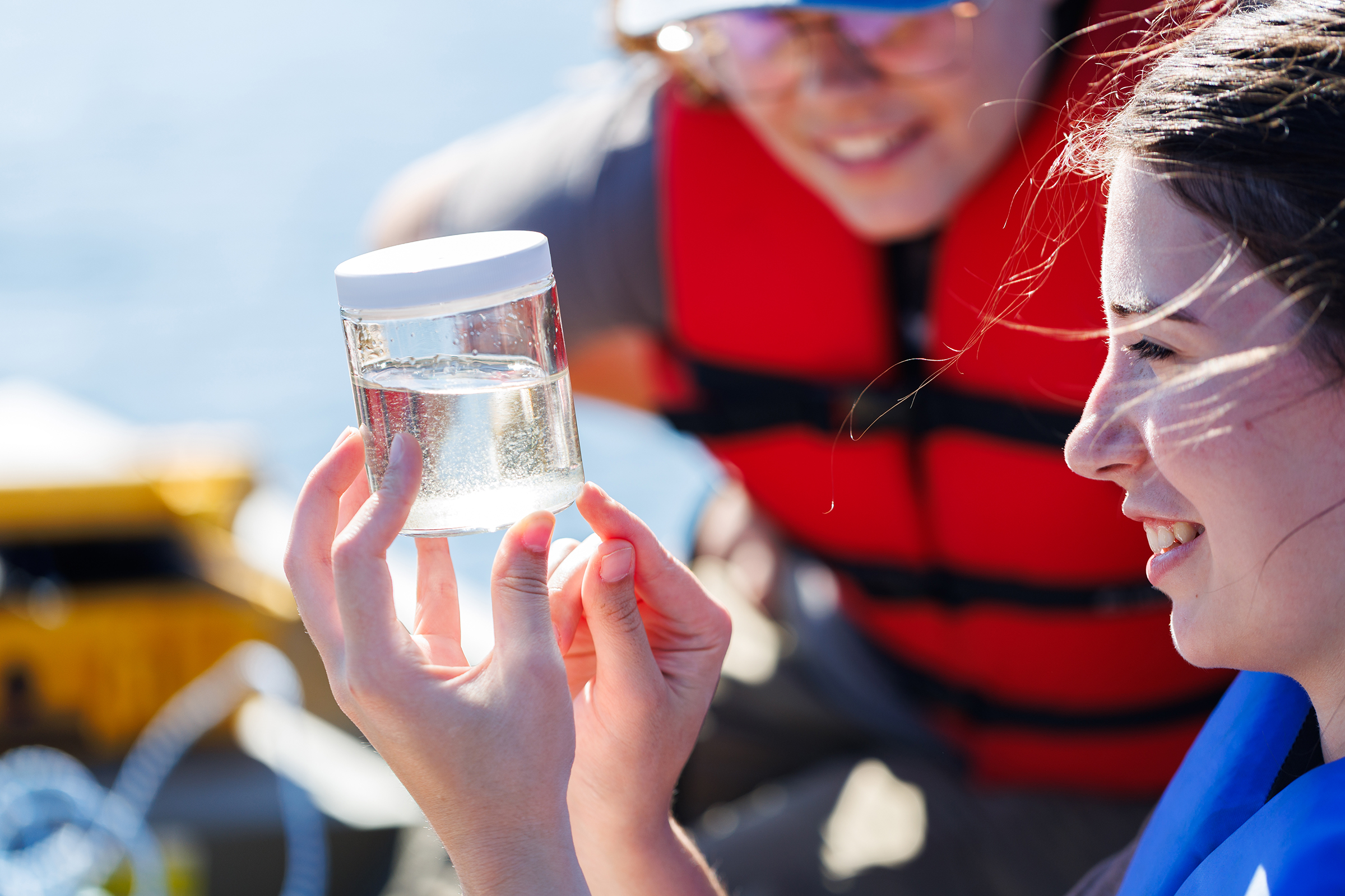 Two individuals in life jackets examine a sample of water in a clear container while on a boat.