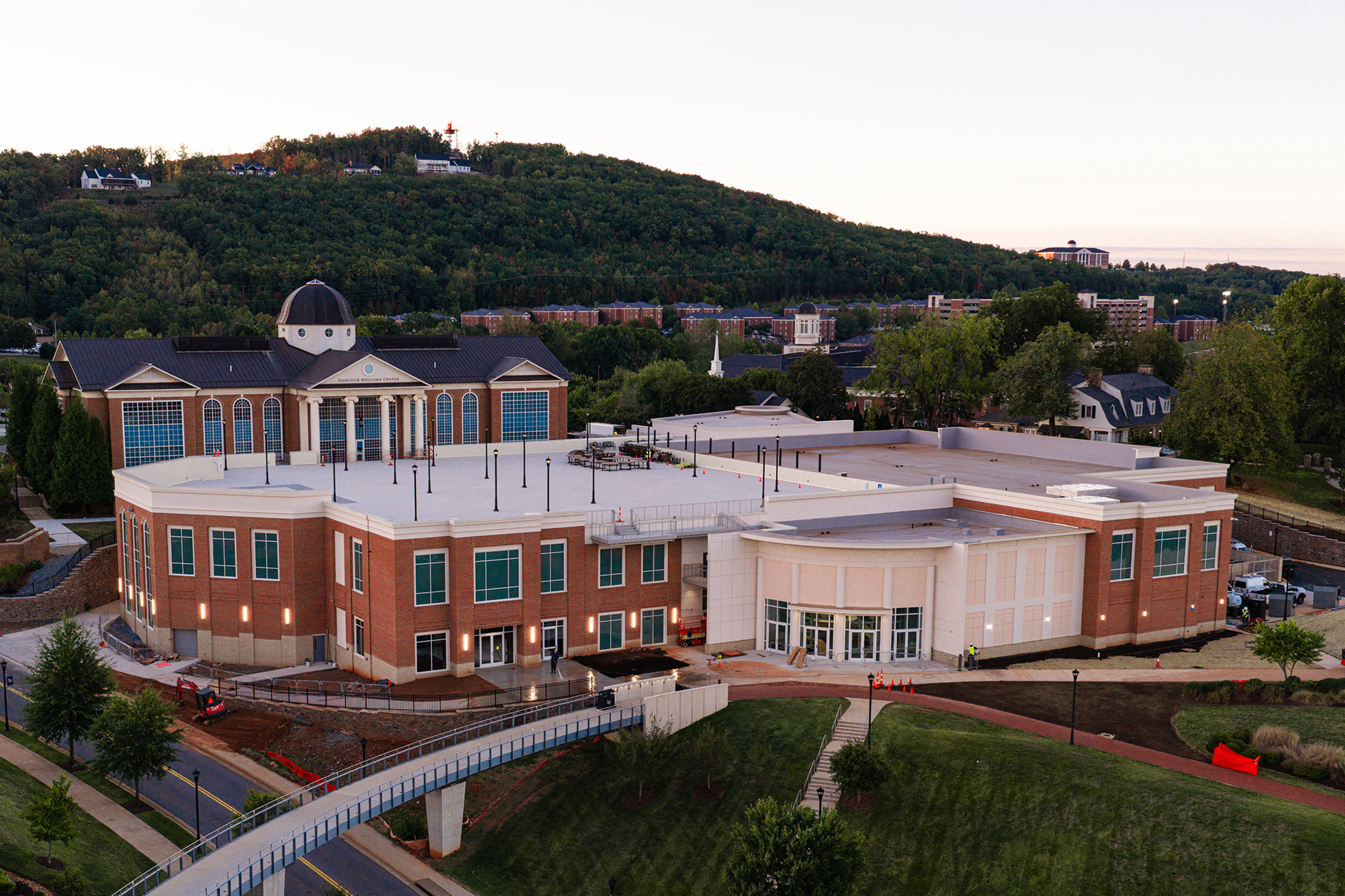 An aerial view of a modern campus building complex with a distinctive dome, surrounded by green hills, pathways, and a pedestrian bridge.