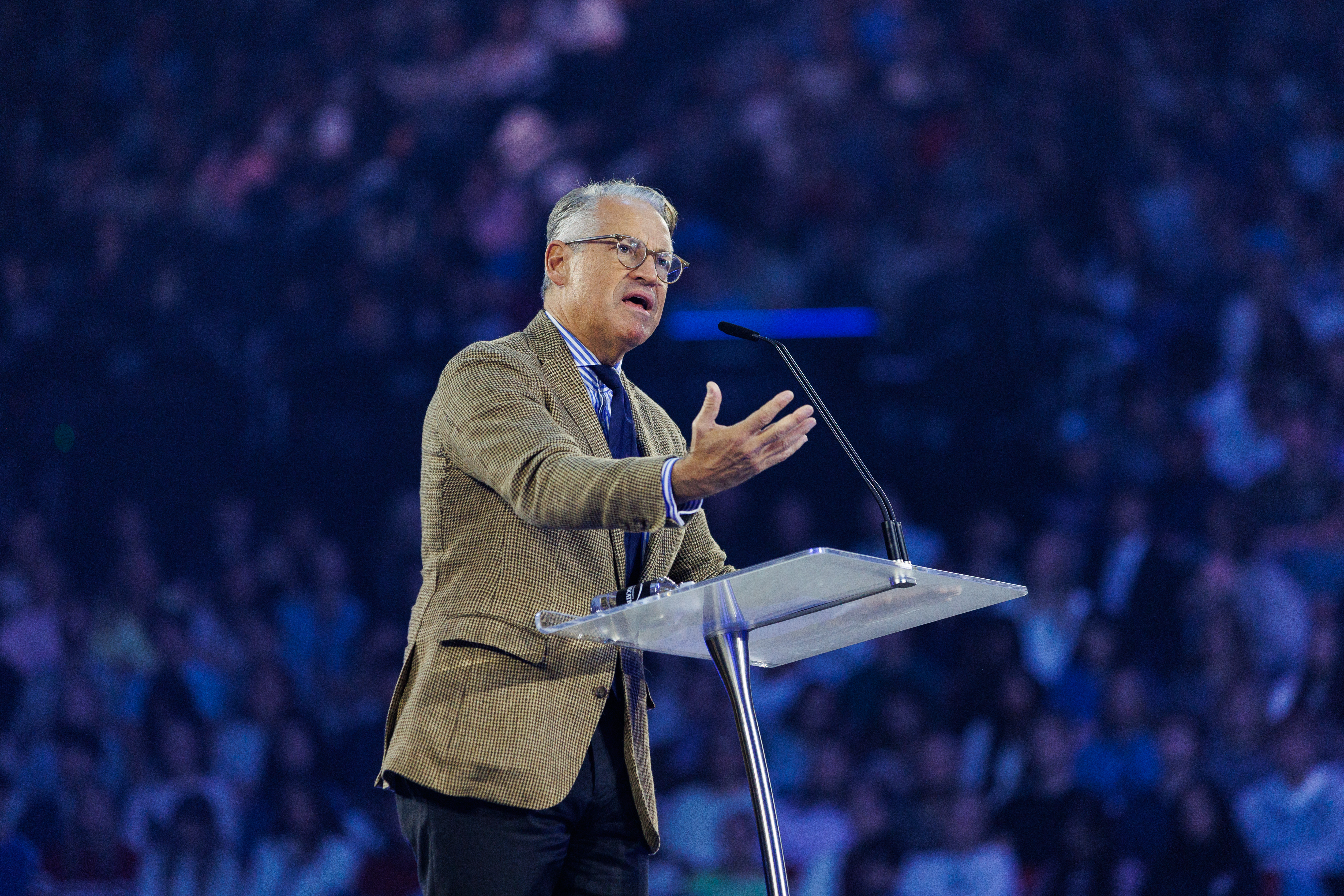 A person in a patterned blazer and glasses stands at a podium, gesturing while speaking to a large, blurred audience under blue lighting.