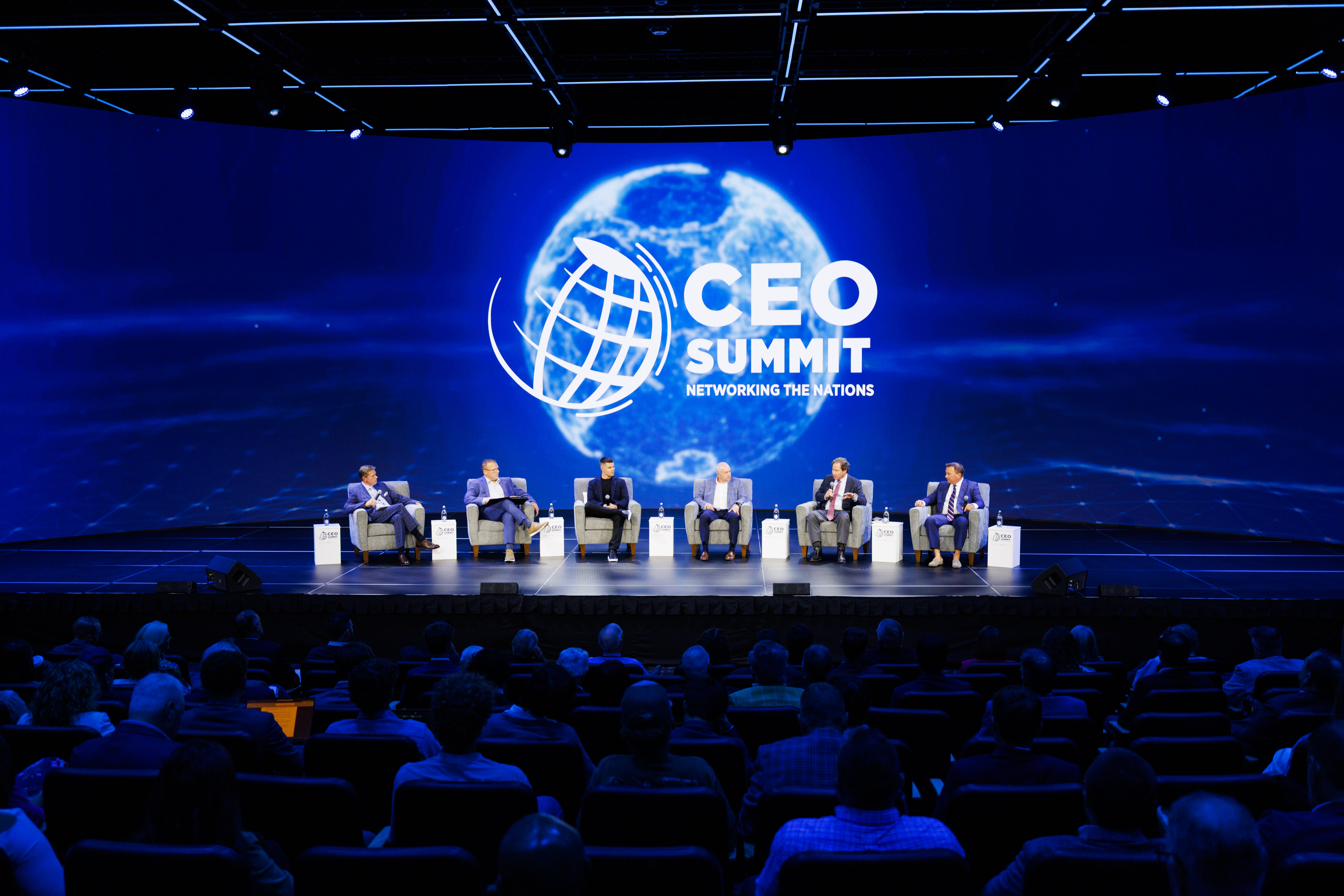 Six individuals sit on a stage at the CEO Summit, with a large blue screen displaying the event logo and a globe behind them, as an audience watches from the darkened auditorium.
