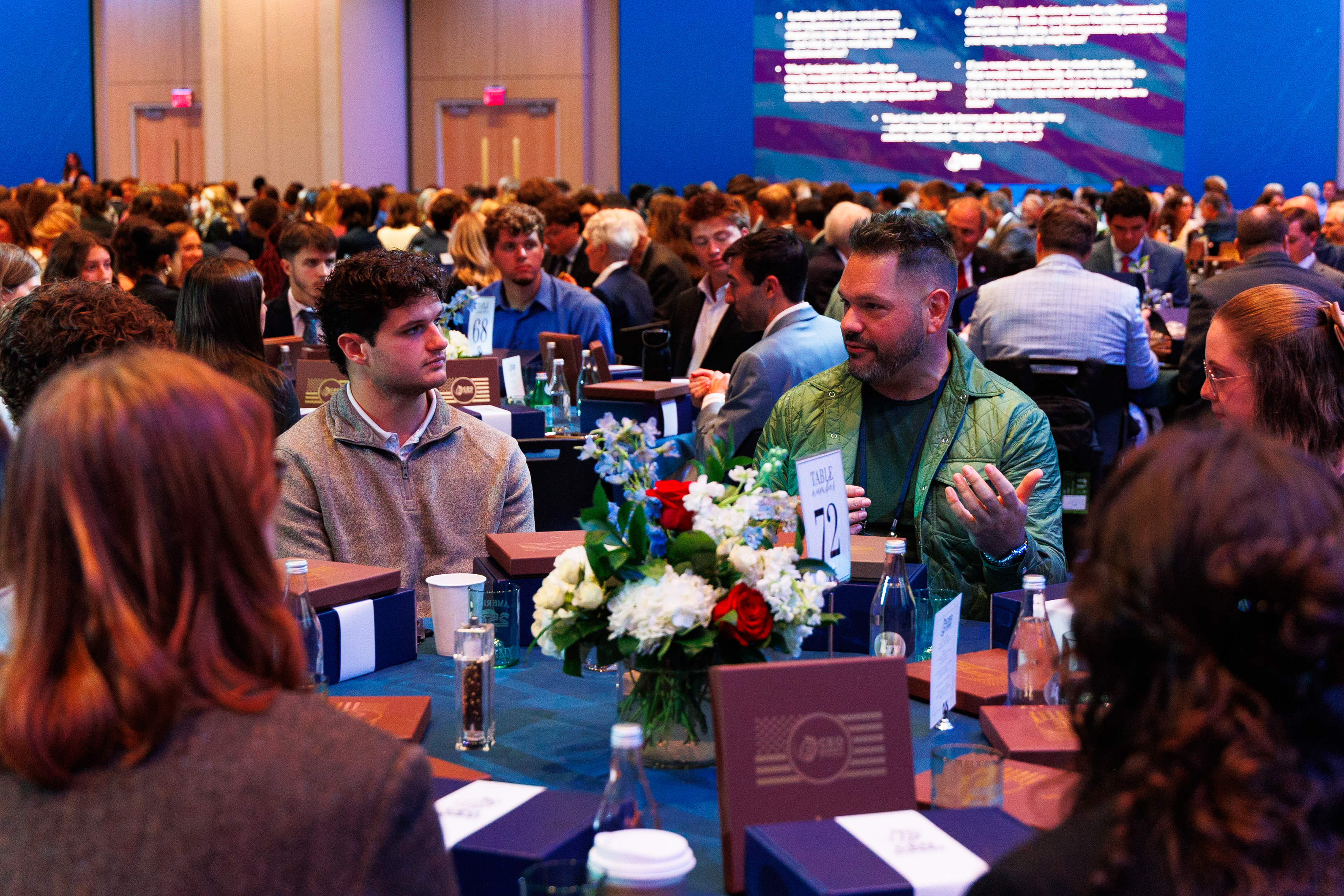 A large gathering of people seated at tables in a conference hall, engaged in conversation and dining, with a floral centerpiece and a presentation screen visible in the background.