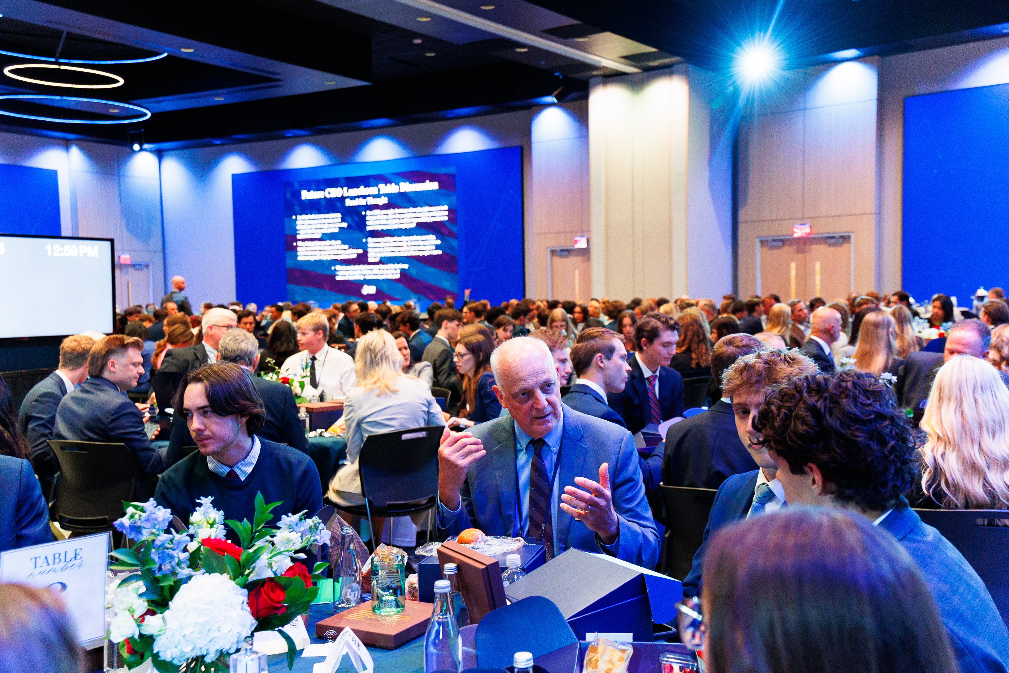 A large conference hall filled with attendees seated at tables, engaged in conversation, with a presentation screen displaying "Future CEO Leadership Role Discussion" in the background.