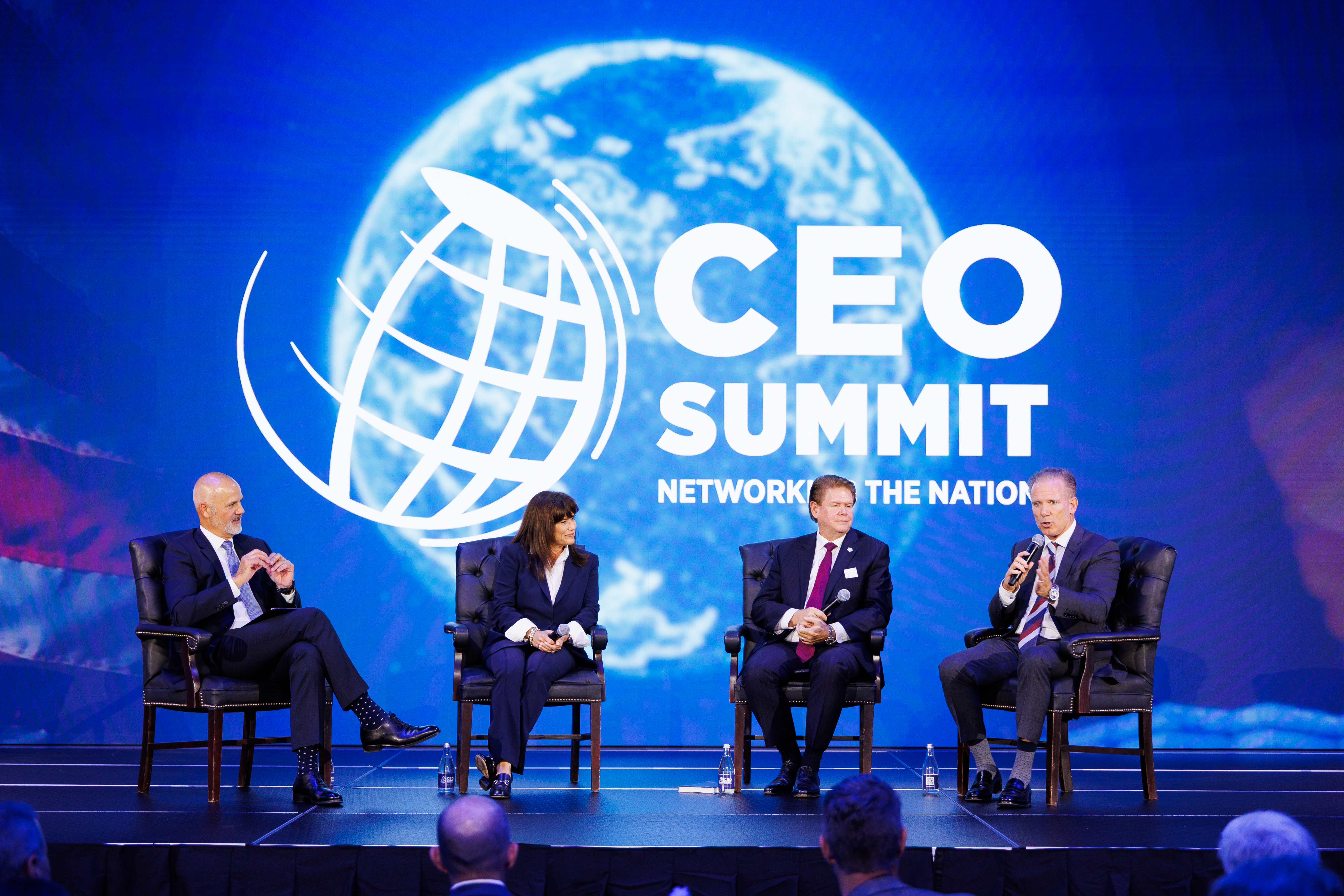 Four individuals participate in a panel discussion at the CEO Summit, seated on stage before a large screen displaying the event's logo and a globe.