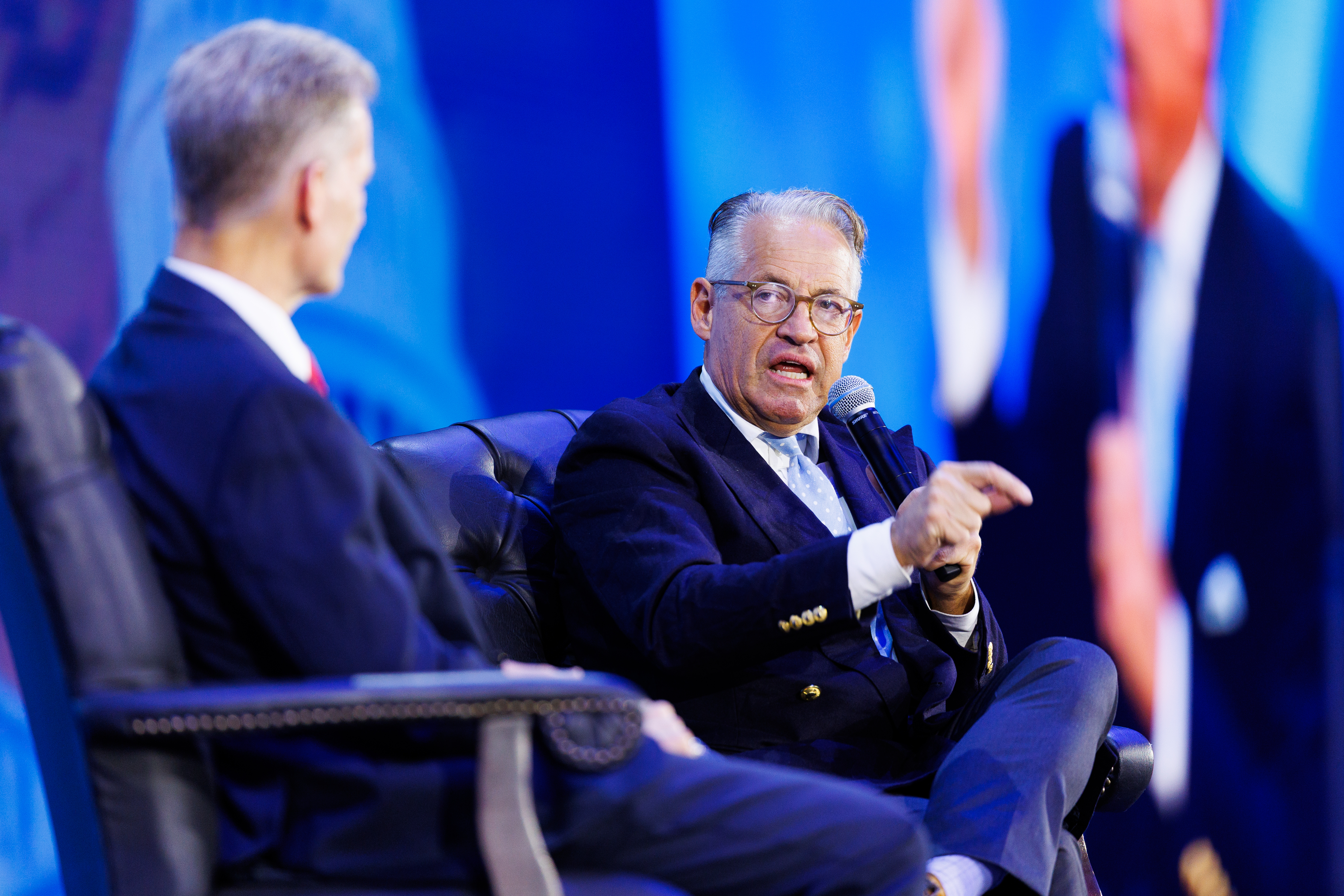 An individual in a suit and glasses speaks into a microphone while seated on a stage, engaging with another person in formal attire.