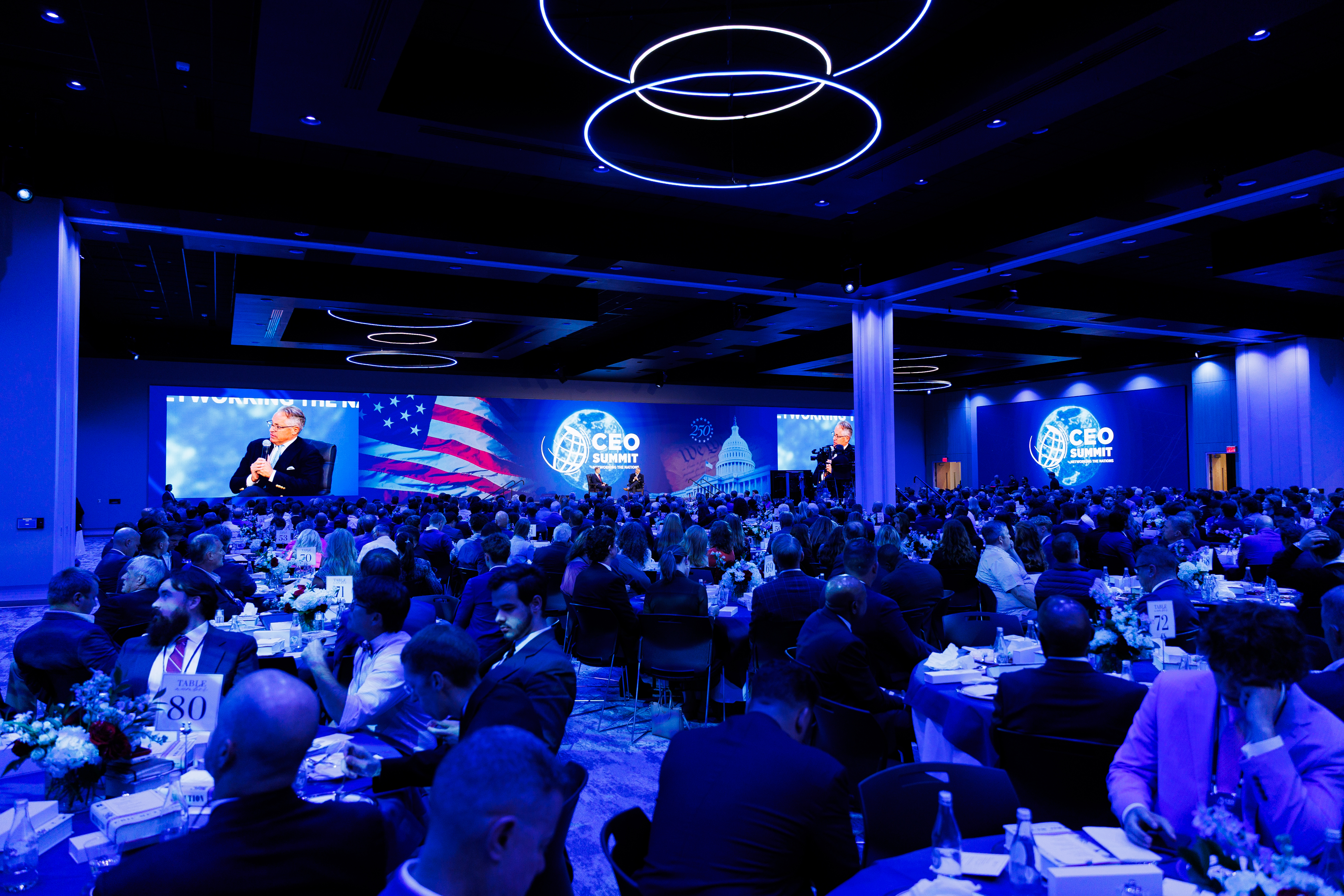 A large conference hall filled with attendees seated at round tables, watching a speaker on a large screen under blue lighting.