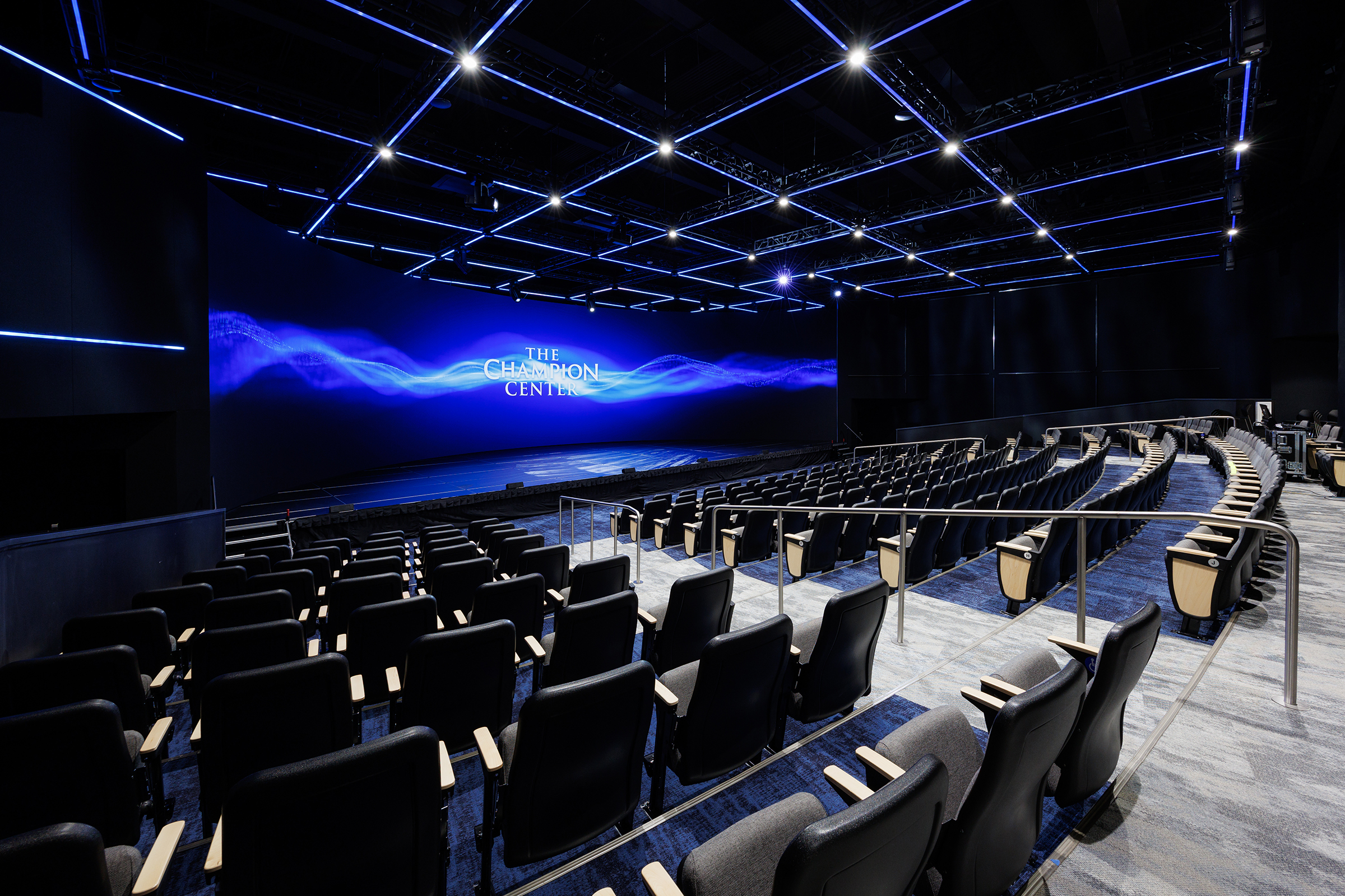 A modern auditorium with black seats and a large blue screen displaying "The Champion Center" under a ceiling with blue lighting.