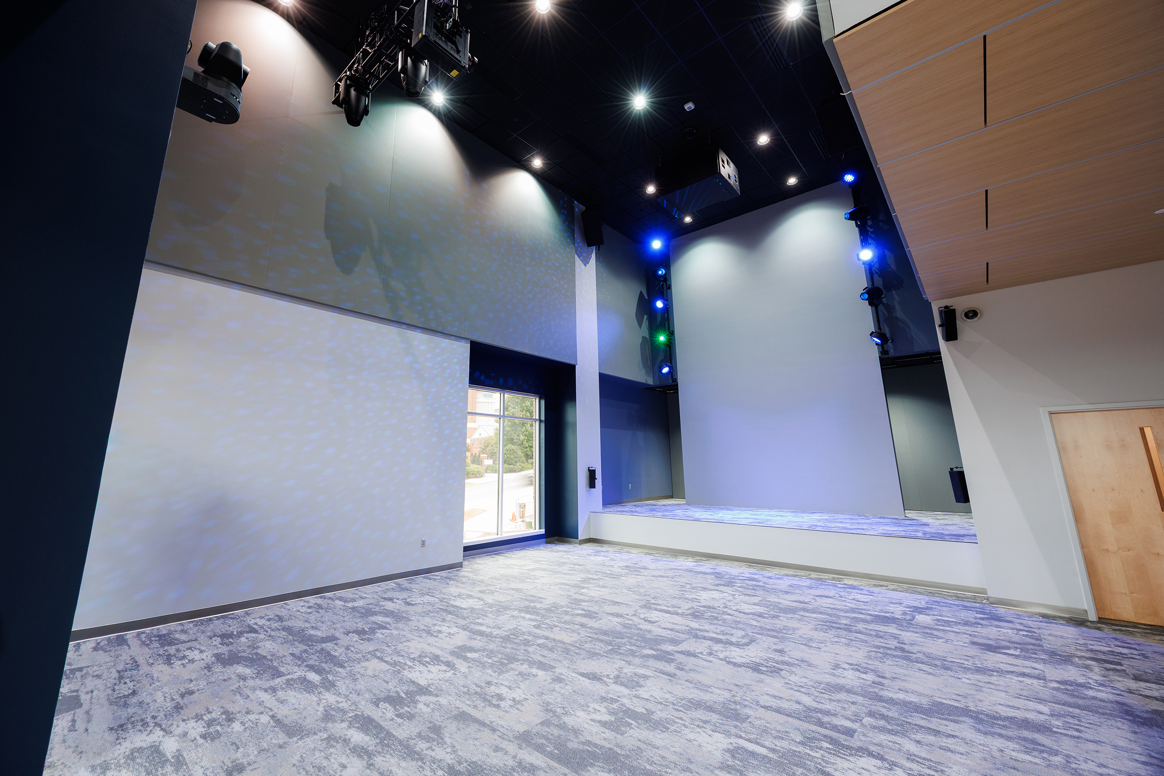 An empty performance venue with a stage, lighting rigs, and a large screen, illuminated by spotlights and colored stage lights.