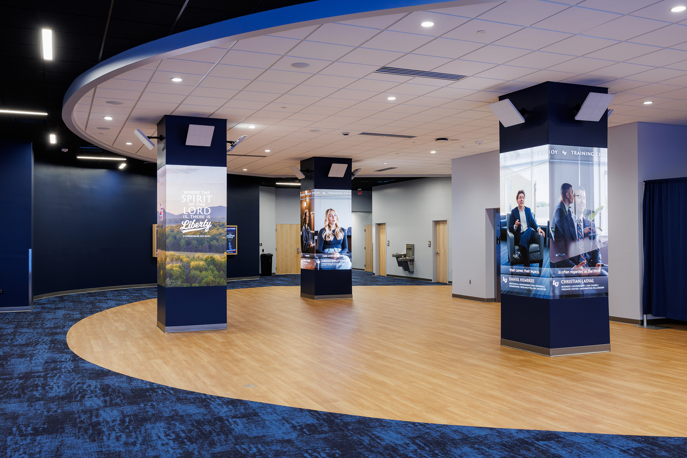 A modern indoor space features three tall pillars with promotional displays, set against a curved wooden floor and blue carpeting under a circular ceiling.
