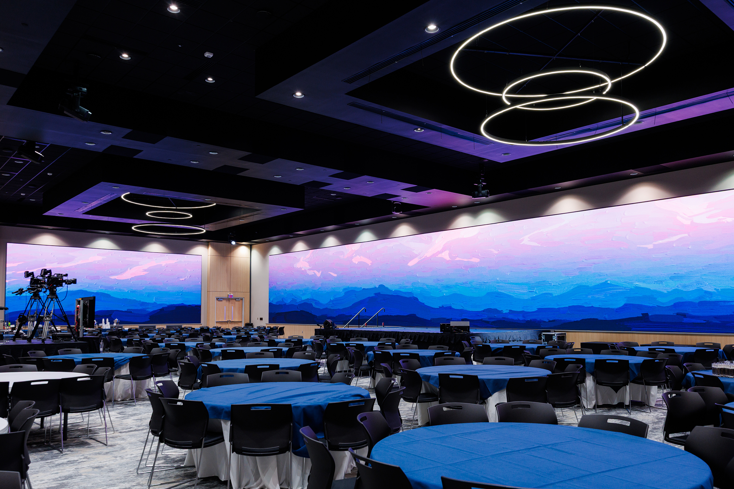 A modern conference hall features round tables with blue tablecloths, black chairs, and large screens displaying a serene mountain landscape under a twilight sky, illuminated by circular pendant lights.