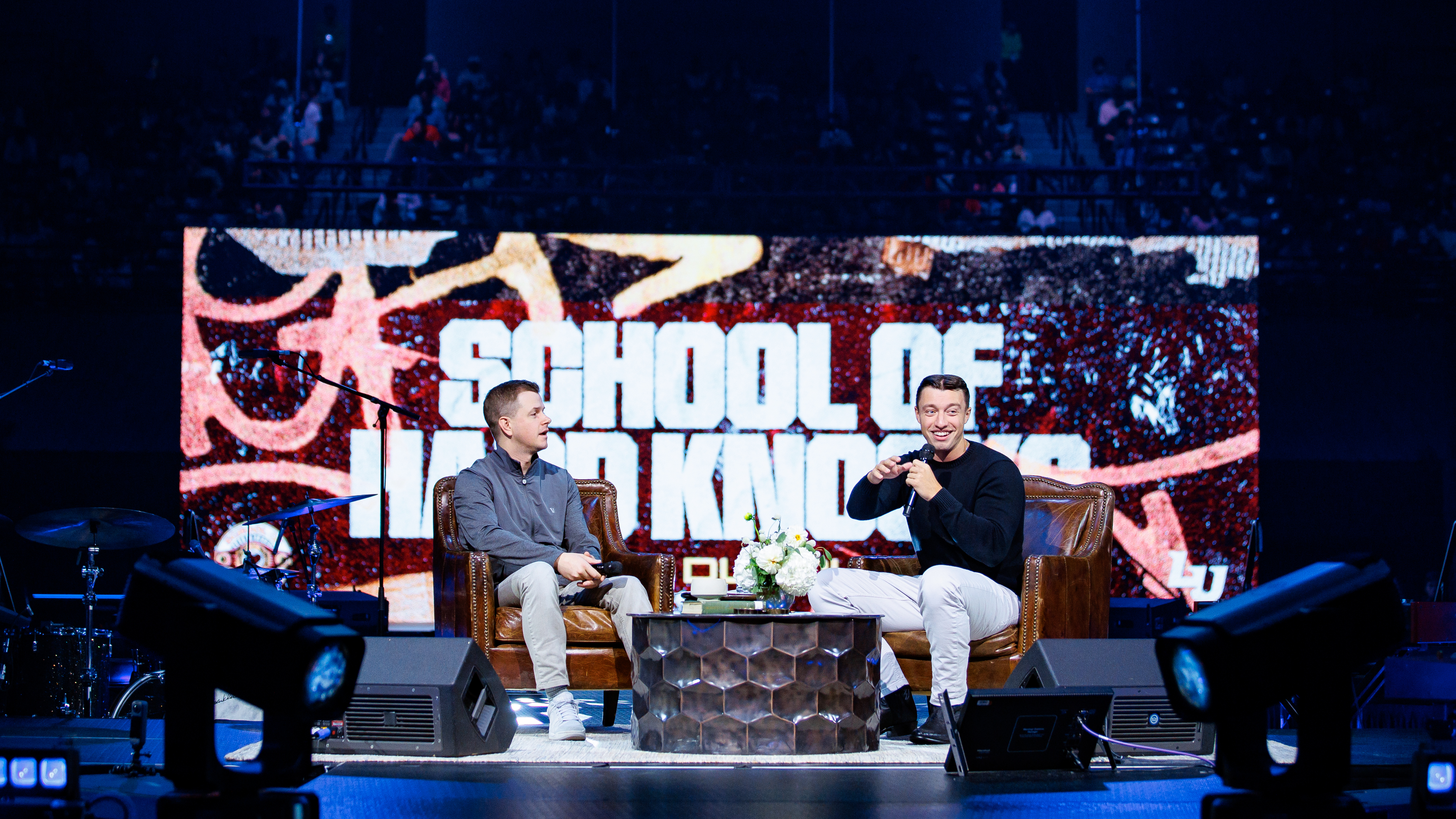 Two individuals sit on a stage in front of a large screen displaying “SCHOOL OF HARD KNOCKS,” engaged in conversation with a small table between them.