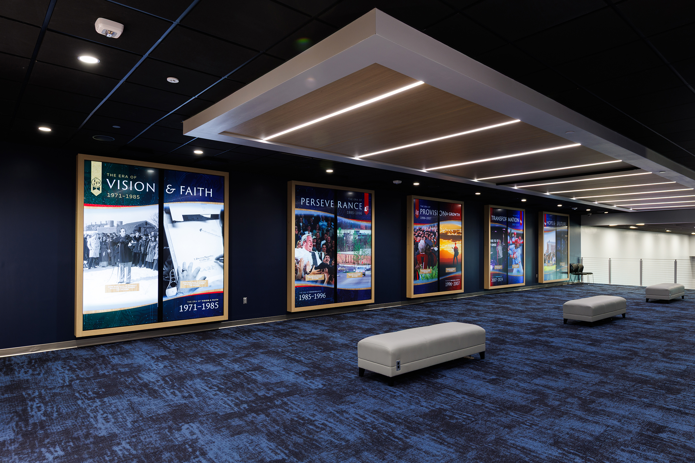 A modern exhibition space features large illuminated displays chronicling historical eras, set against dark walls and blue carpeting with minimalist seating.