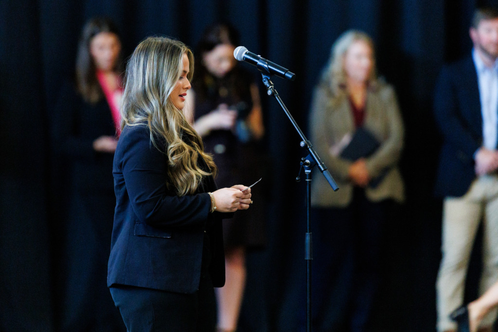 A person in a dark suit stands at a microphone, speaking to an audience while holding a small object, with blurred figures in the background.