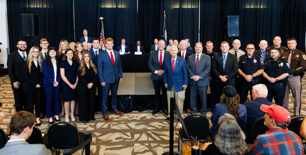 A group of individuals, including officials and staff, pose together in a formal setting with flags and a podium.
