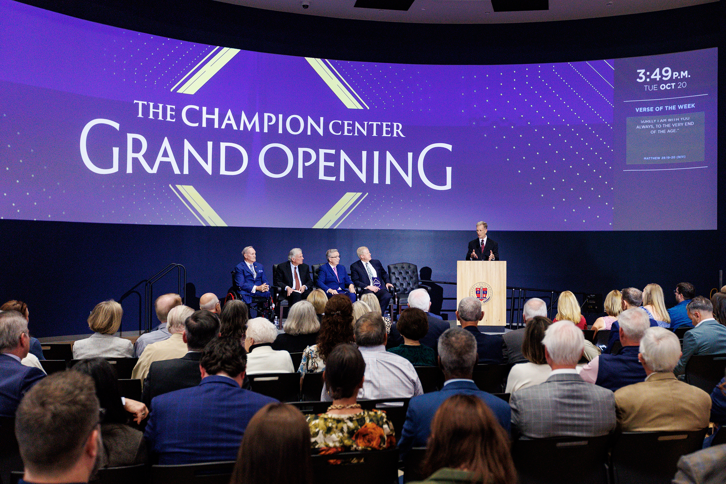 A speaker addresses an audience at the grand opening of The Champion Center, with a large screen displaying the event name and date.