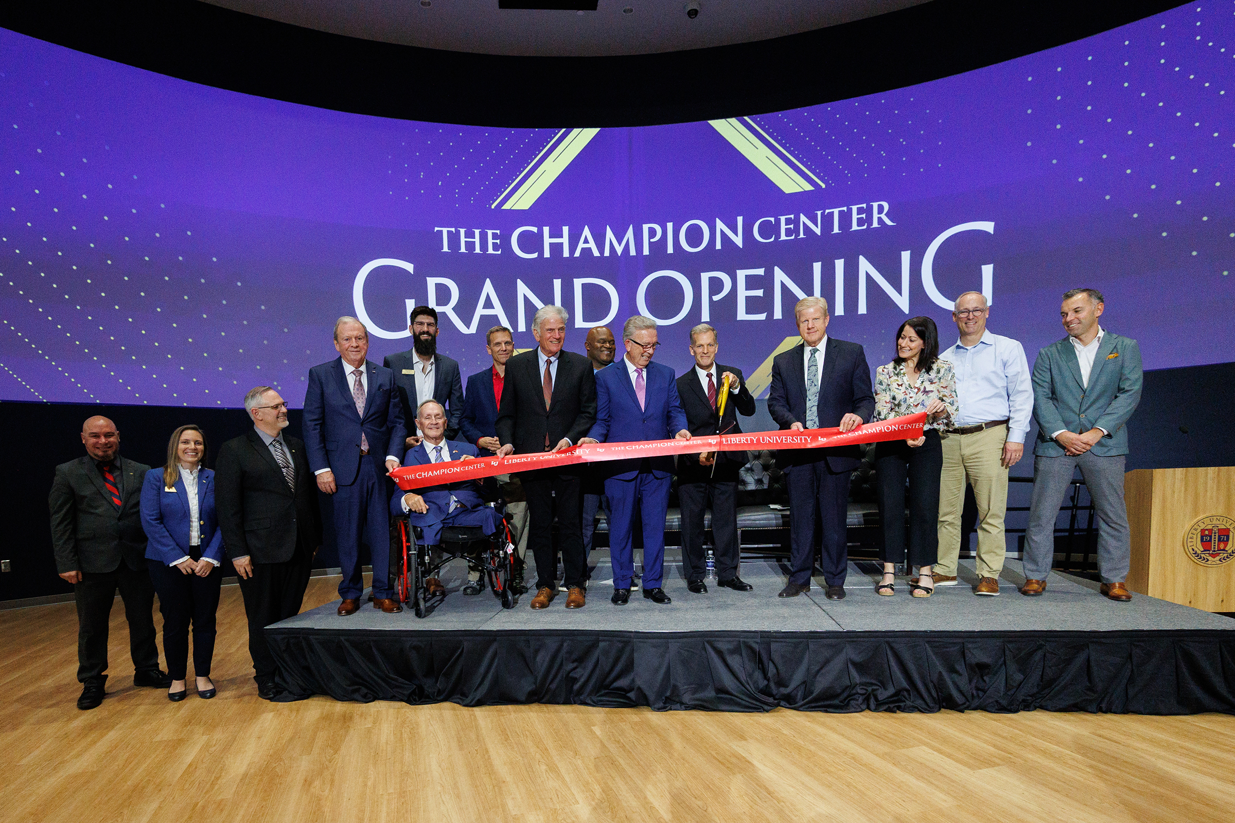 A group of people, including one in a wheelchair, stand on a stage cutting a red ribbon during the grand opening of The Champion Center.