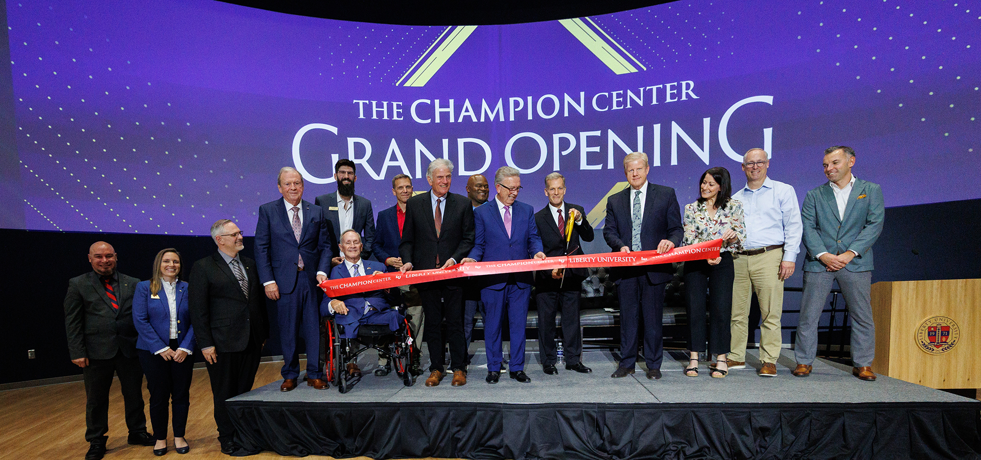 A group of people, including one in a wheelchair, stand on a stage cutting a red ribbon during the grand opening of The Champion Center.