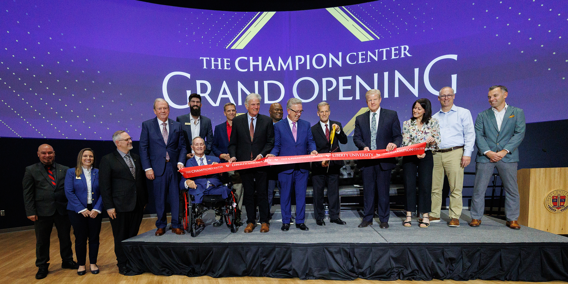 A group of people, including one in a wheelchair, stand on a stage cutting a red ribbon during the grand opening of The Champion Center.